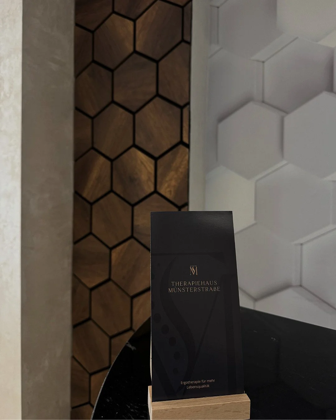 Schwarze Broschüre mit Goldschrift auf einem schwarzen Tisch, im Hintergrund eine Wand mit Holz- und weißen Fliesen im Hexagonmuster.