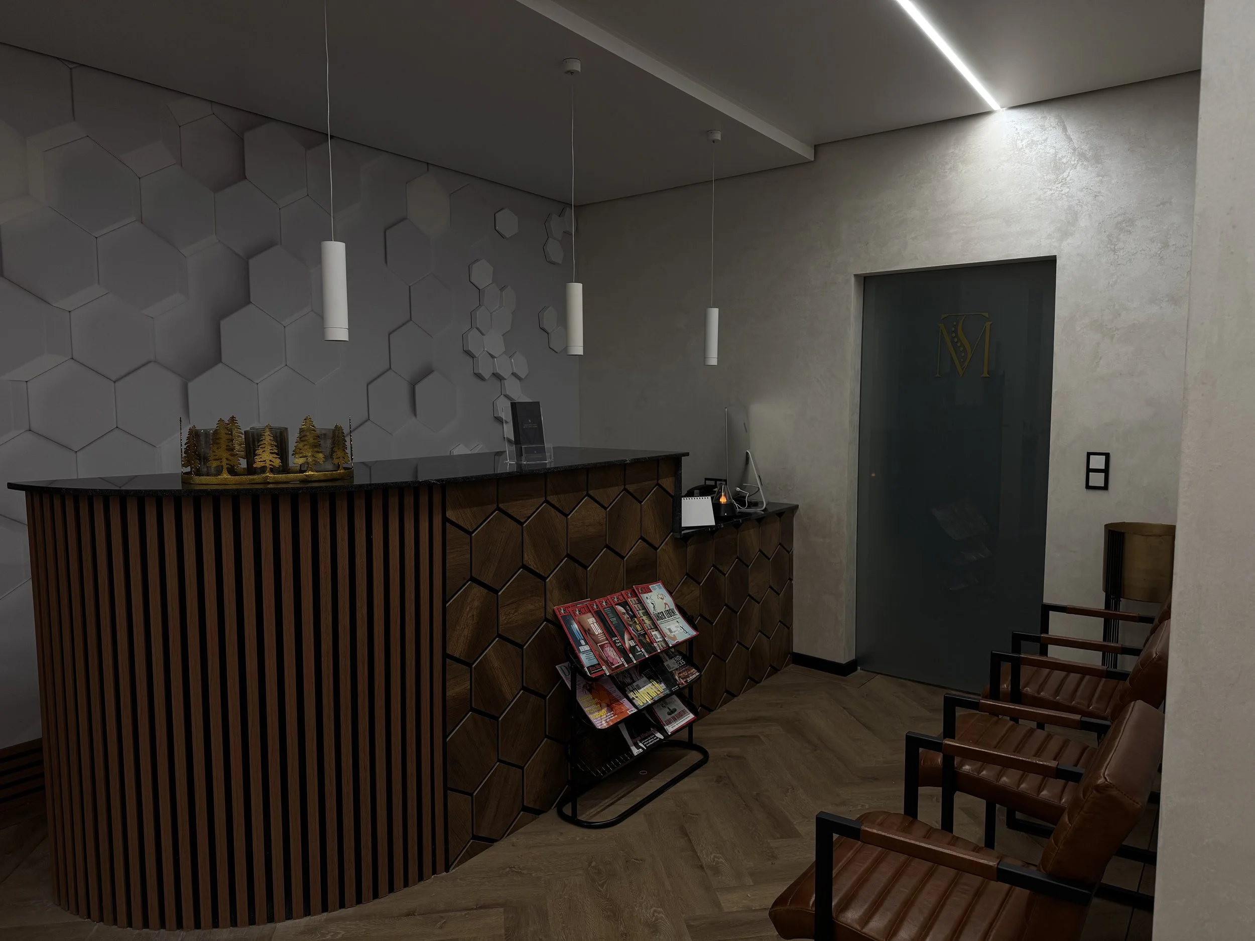Hotelrezeption mit holzvertäfeltem Empfangstresen, grauem Hexagon-Wand hinter dem Tresen, drei weißen Pendelleuchten, Zeitschriftenständer, Computer und Telefon auf dem Tresen, dunkler Tür mit goldenem Logo, braune Ledersessel, Holzfußboden.
