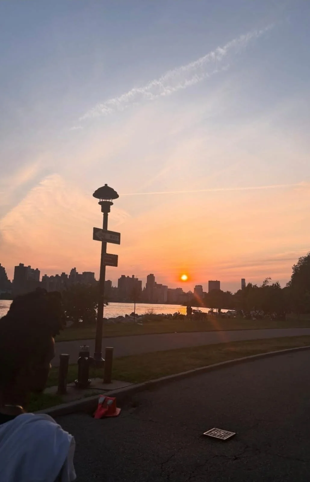 Sunset over a city skyline with some trees and a river in the foreground, a streetlamp, and a person seated near the sidewalk.