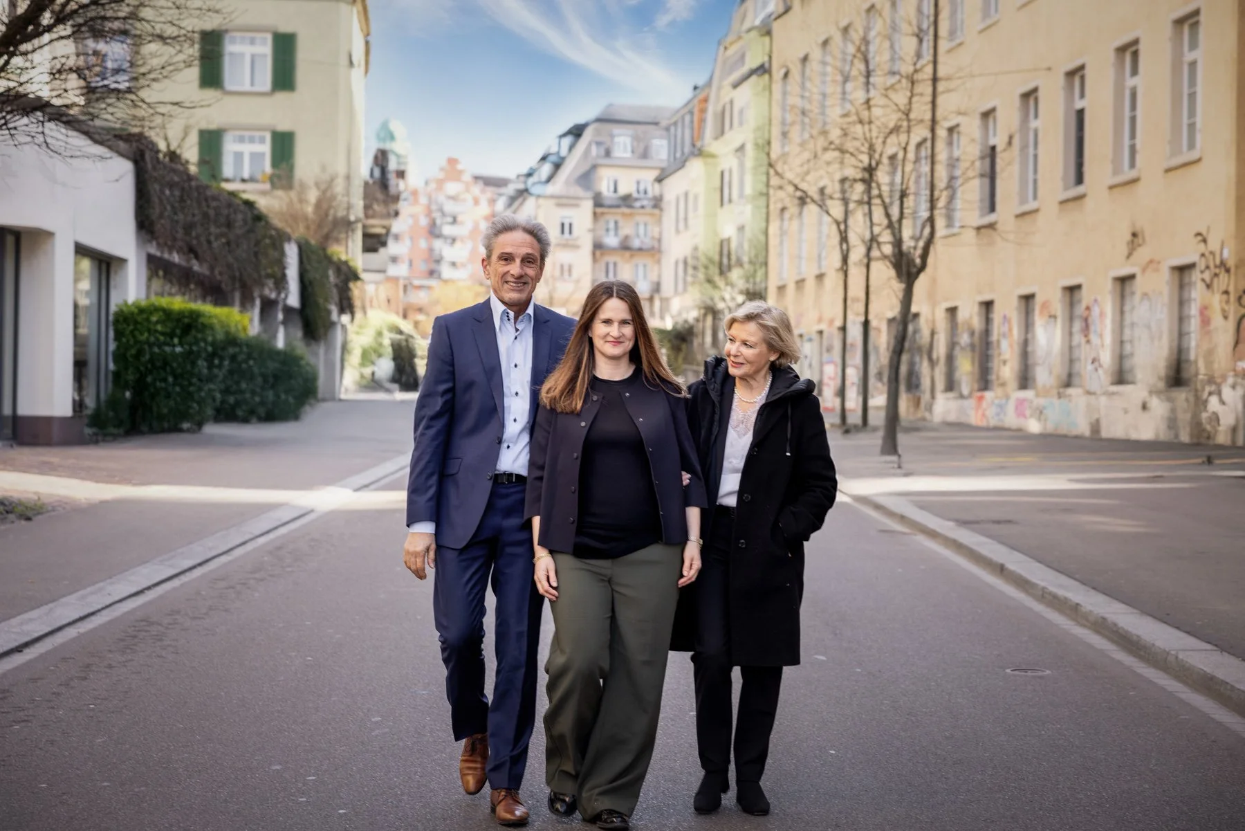 Es handelt sich um das Team von refliktiv Immobilien AG, zwei Frauen und einen Mann, die nebeneinander gehen und lächeln. Im Hintergrund sind mehrstöckige Gebäude mit Graffiti an den Wänden, einige Bäume ohne Blätter und ein blauer Himmel mit Wolken.