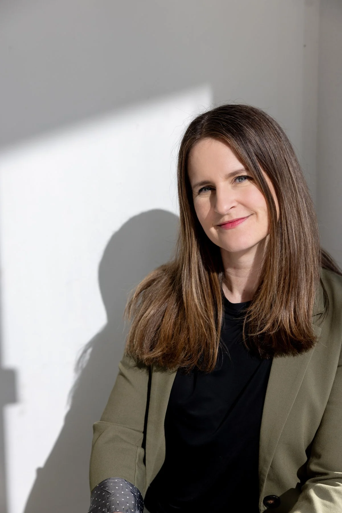 Frau mit braunen, glatten Haaren, lachendem Gesicht, dunkelgrüner Jacke, schwarzem Top, sitzend vor einer weißen Wand mit Schatten.