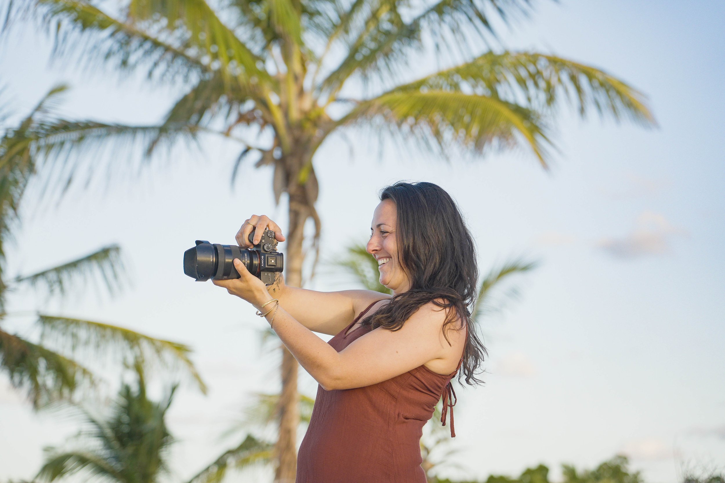 Femme souriante avec appareil photo en plein air, palmiers en arrière-plan, ciel clair.
