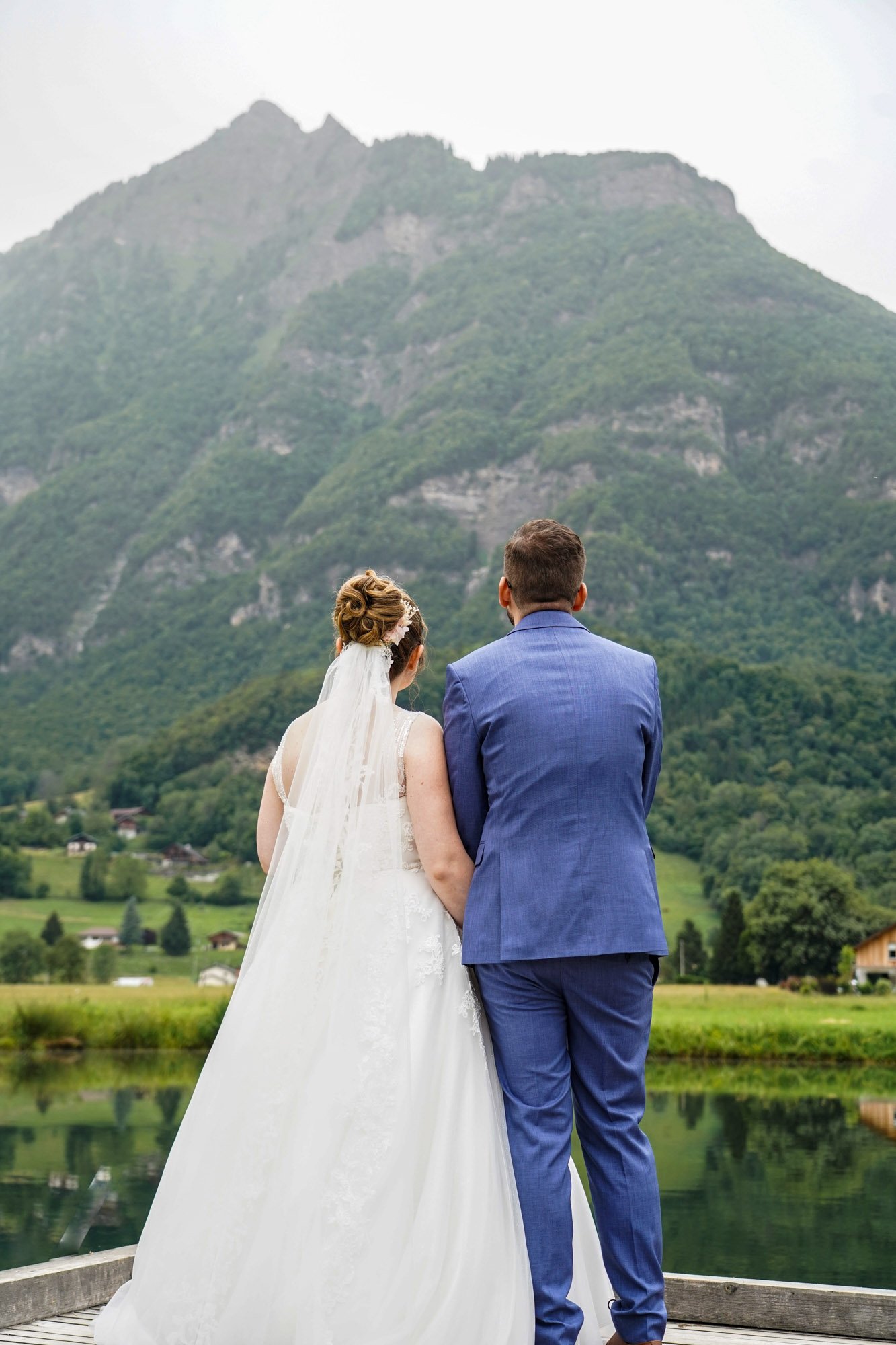 Un couple marié, vu de dos, regarde un paysage montagnard avec un lac et des maisons dans la vallée.
