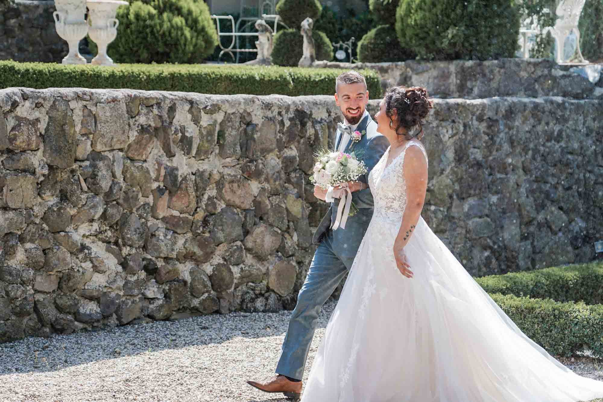 Couple de mariés souriants marchant dans un jardin avec un mur en pierre et des sculptures en arrière-plan.