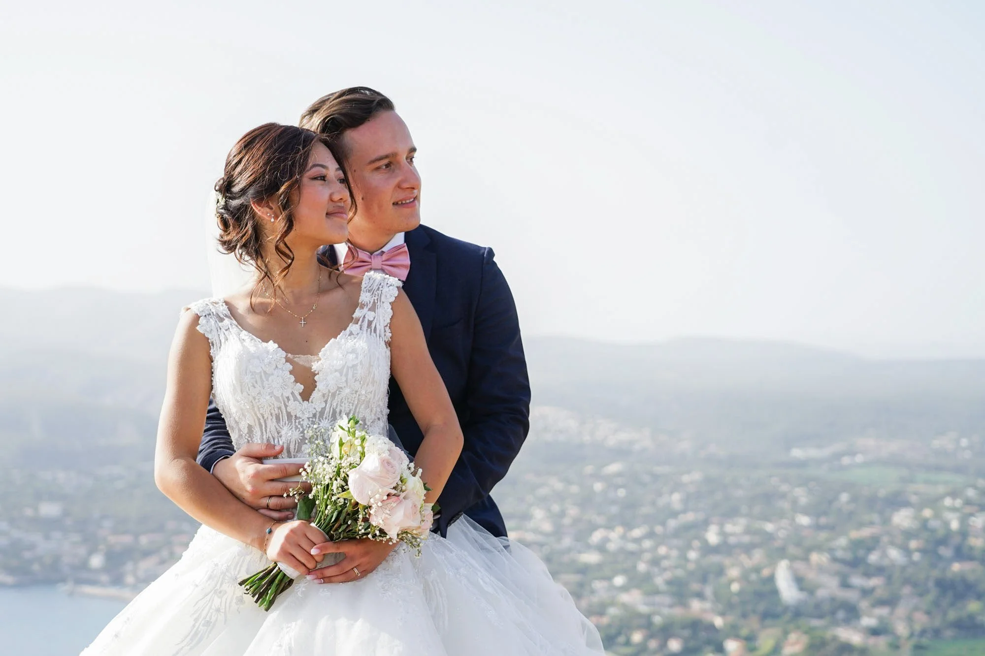 Un couple de mariés pose en plein air avec une vue panoramique en arrière-plan, la mariée tient un bouquet de fleurs.
