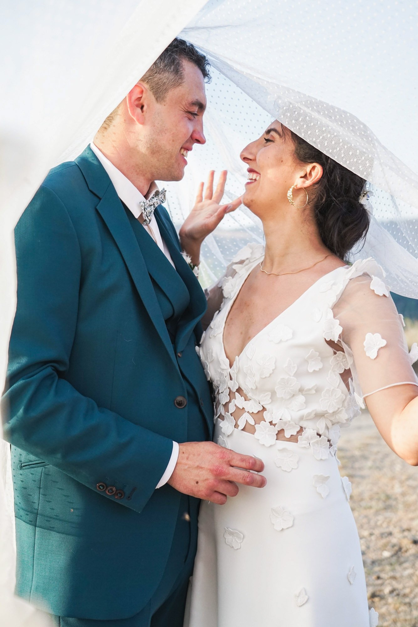 Couple de mariés souriant sous un voile se regardant avec affection.