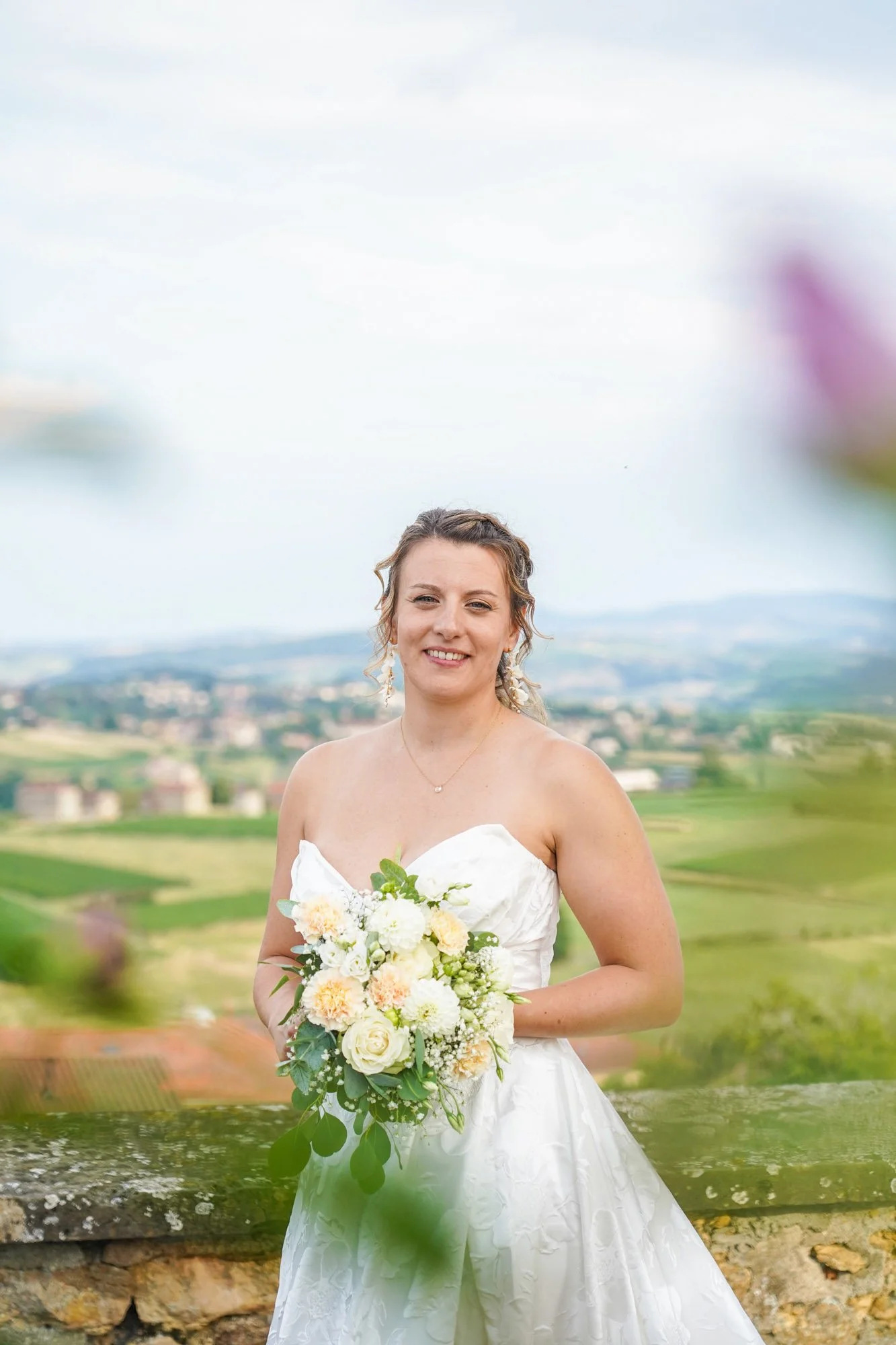 Femme en robe de mariée tenant un bouquet de fleurs dans un paysage rural.