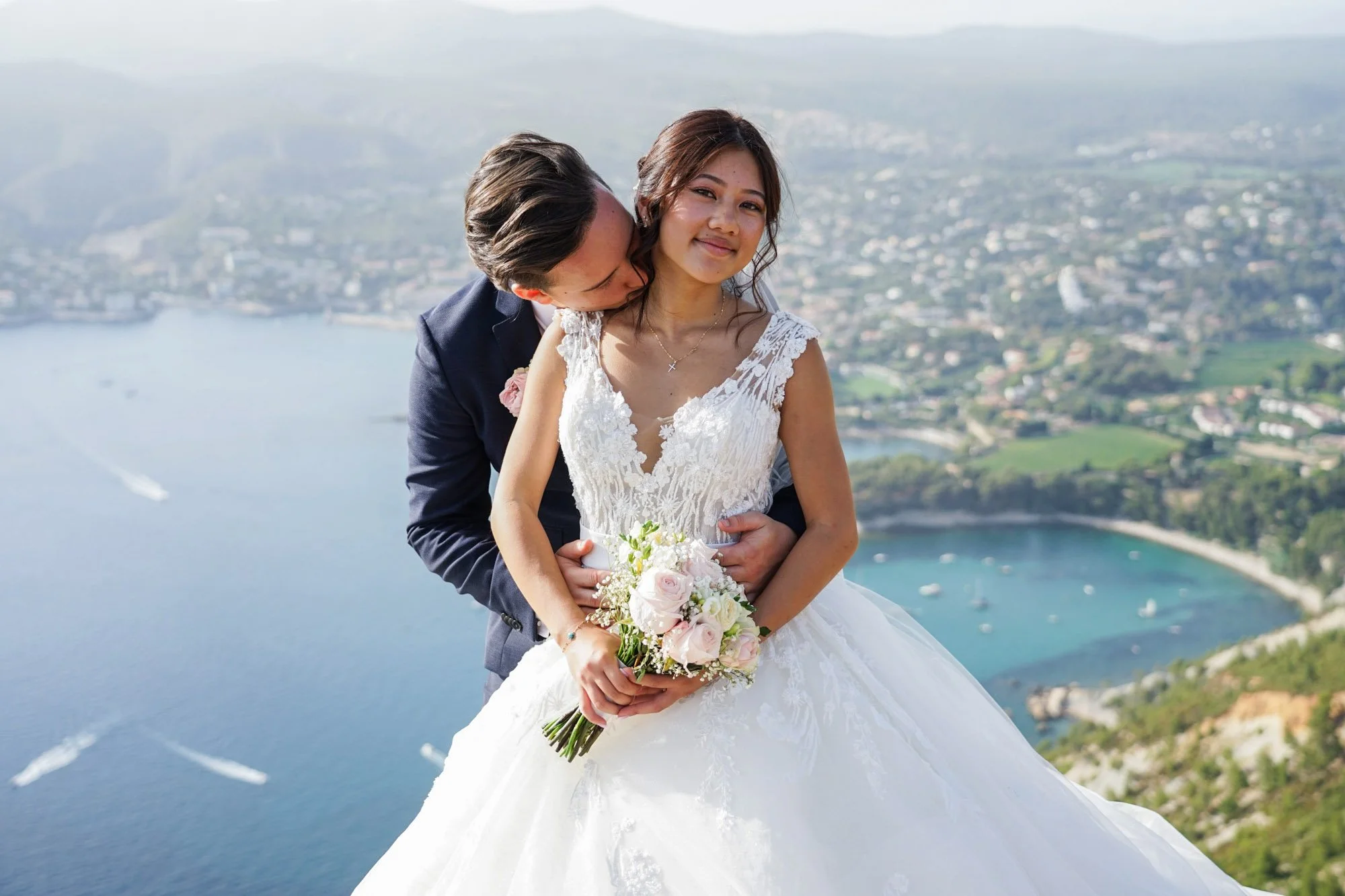 Portrait de mariés, femme avec robe blanche et tenant un bouquet de fleurs roses, avec une vue panoramique sur la mer.