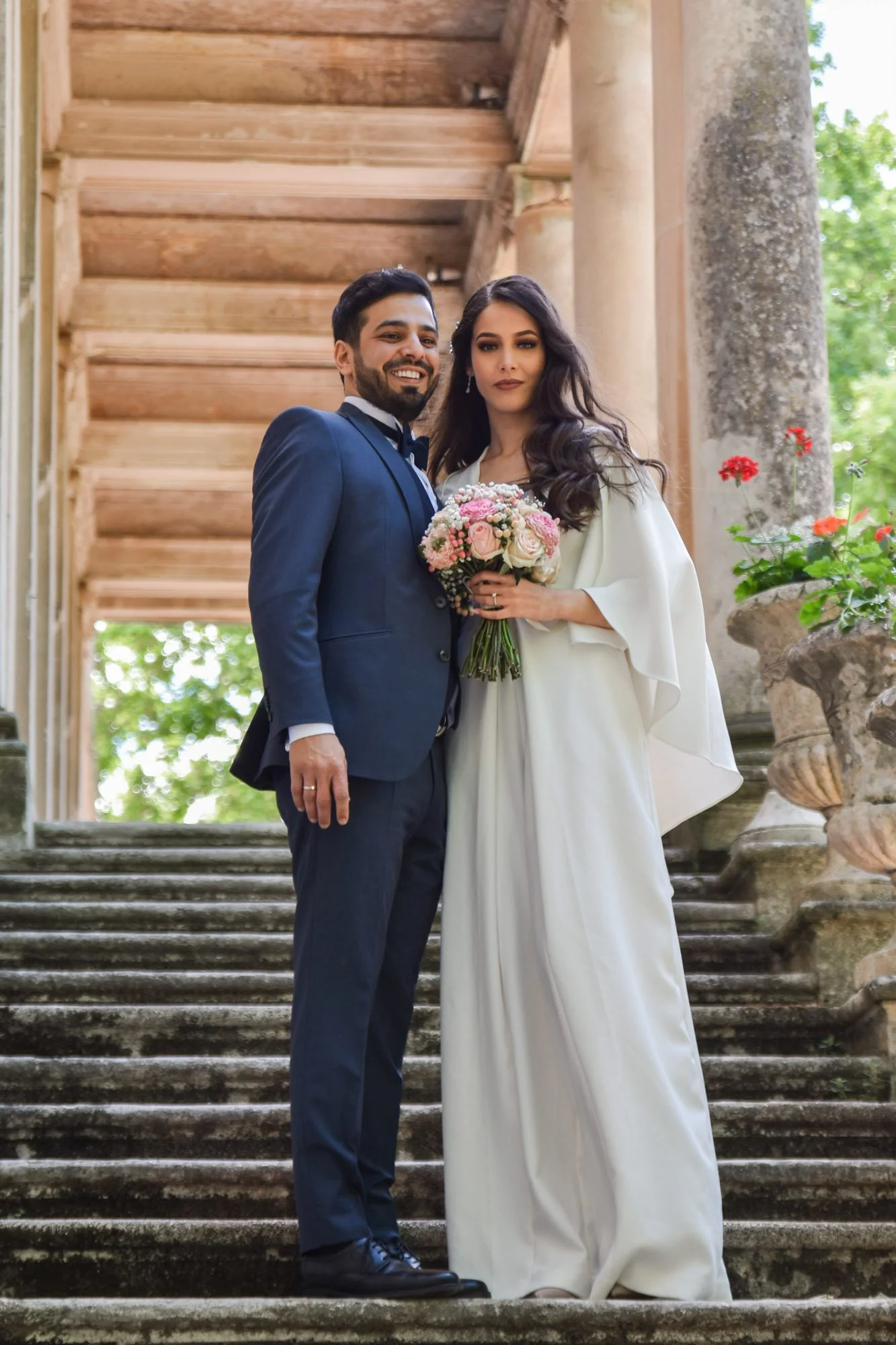 Mariés souriants au photographe sur marches en pierre devant bâtiment historique.