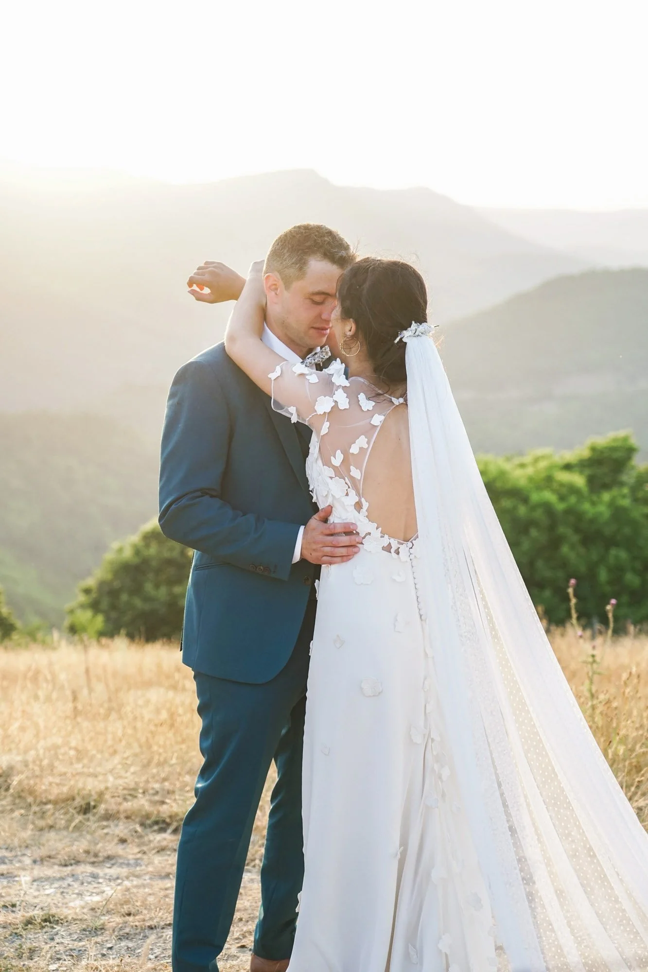 Couple de mariés dans un champ avec des montagnes en arrière-plan au coucher du soleil.