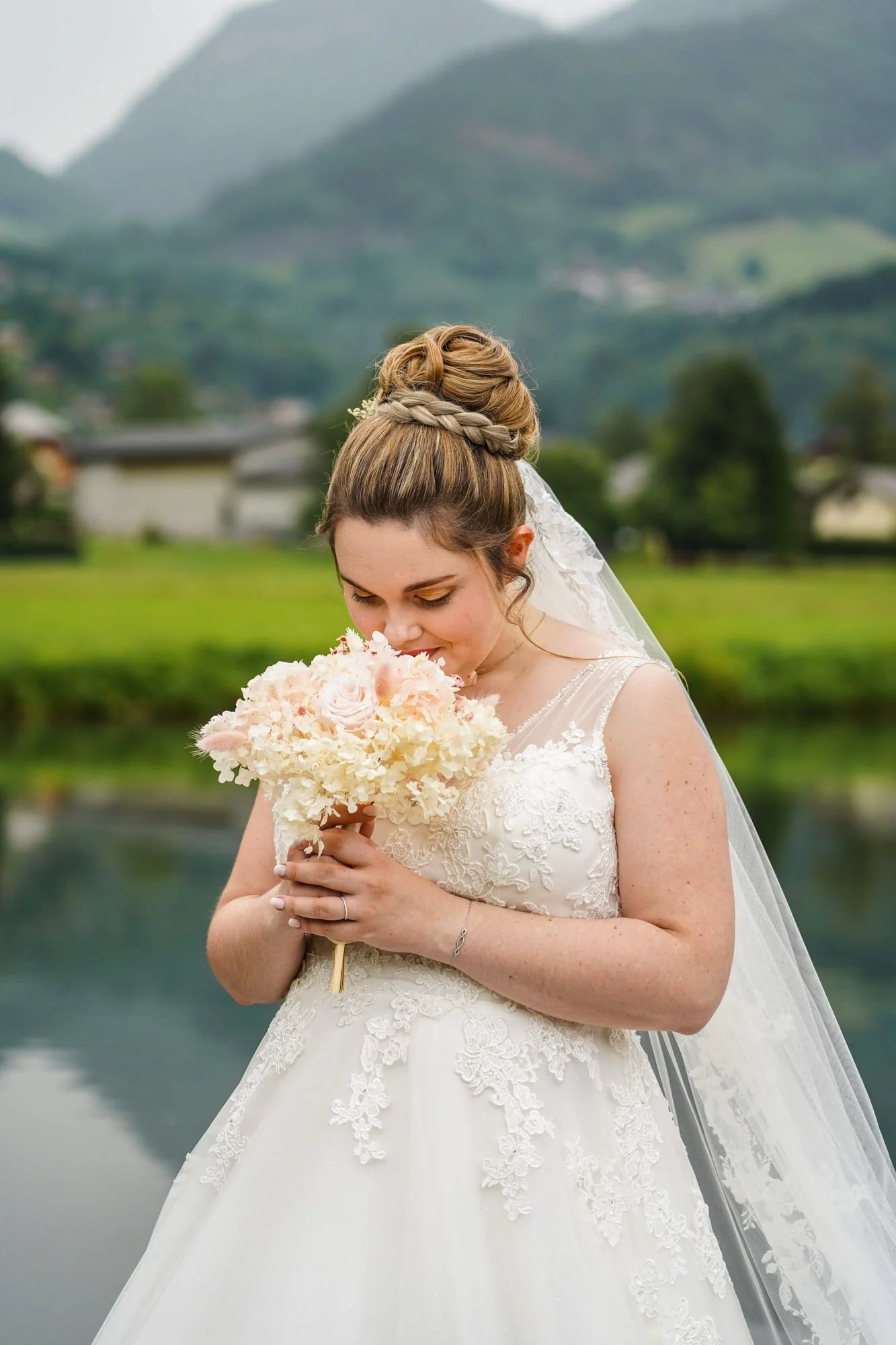 Portrait mariée robe blanche avec bouquet de fleurs séchées. Paysage naturel avec eau, collines verdoyantes et montagnes en arrière-plan.