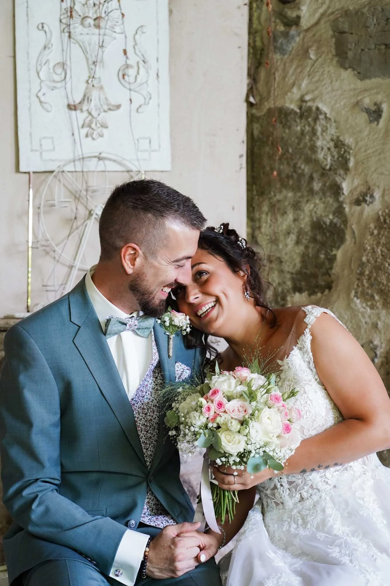 Portrait mariage couple souriant yeux dans les yeux. Bouquet de fleurs roses et blanches, costume marié bleu canard, intérieur rustique.