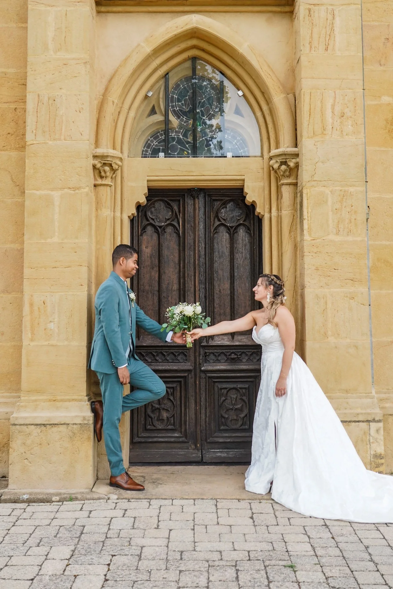 Couple de mariés devant porte en bois ancienne, la mariée en robe blanche et le marié en costume bleu, échangeant un bouquet de fleurs.