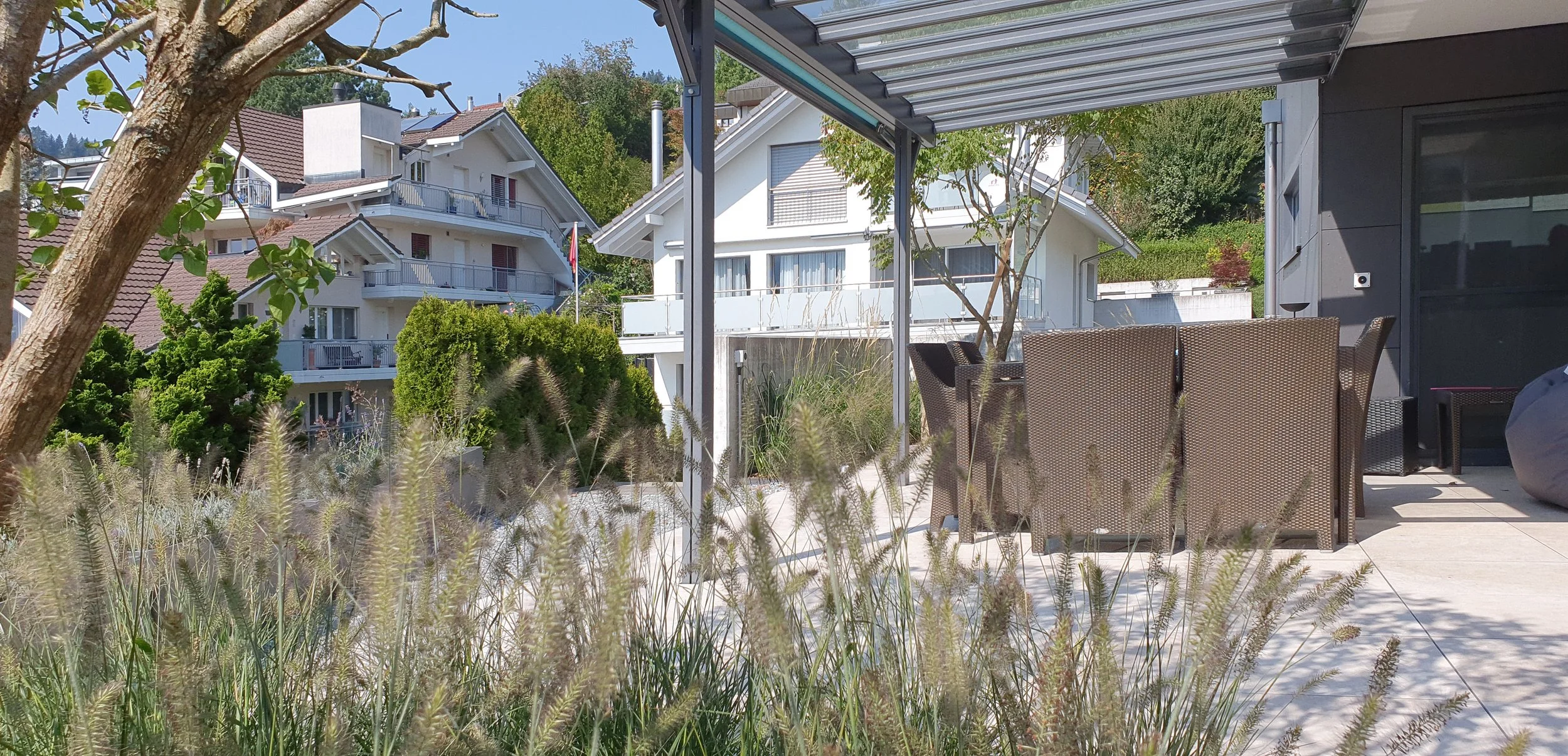 Blick auf eine moderne Terrasse mit Gartenmöbeln, umgeben von Pflanzen und Bäumen, mit mehreren weißen Häusern im Hintergrund.