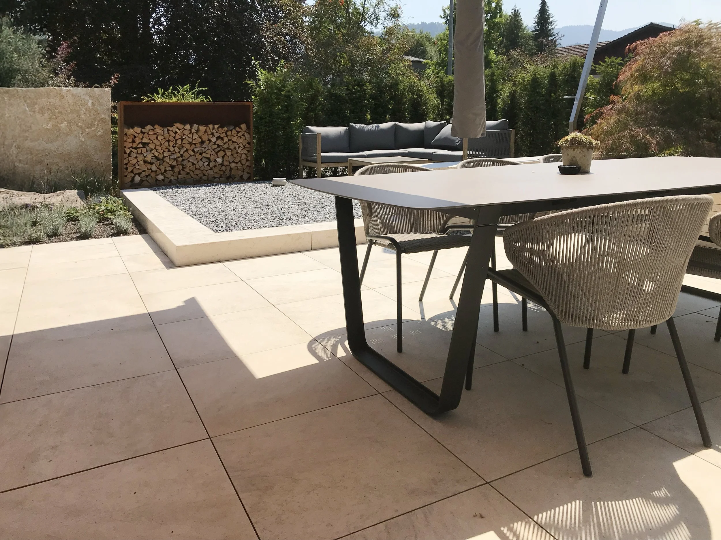 Ein Außenbereich mit Gartenmöbeln, einem Holzaufbewahrungsraum für Feuerholz, und einer Terrasse mit Fliesen, umgeben von Bäumen und Hügeln im Hintergrund.