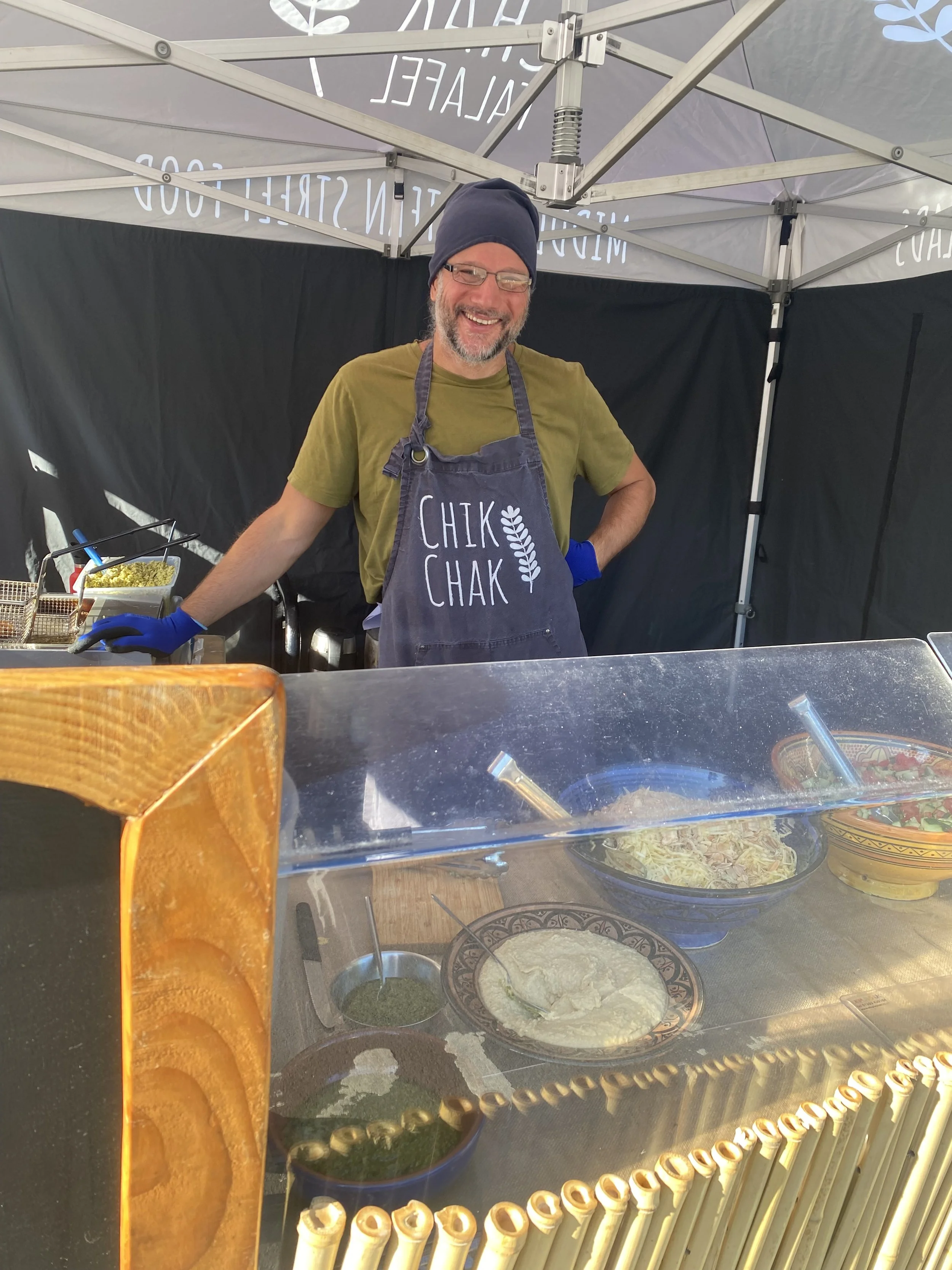 A smiling man wearing glasses, a navy beanie, a green t-shirt, an apron with the words 'Chik Chak' and a leaf design, standing inside a food stall with a black canopy. The stall displays various bowls of food and ingredients, including pasta, herbs, and salads.