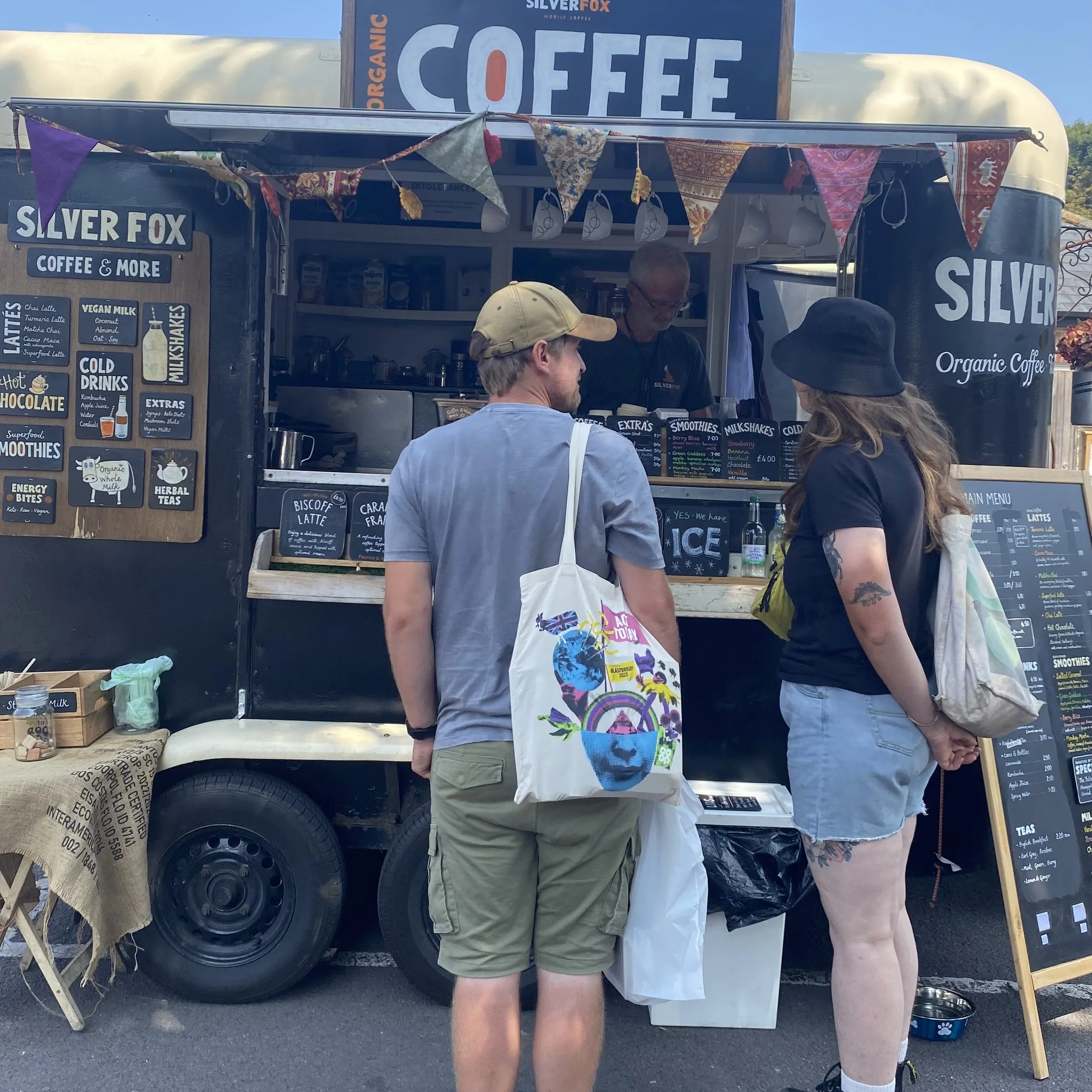 People standing at a coffee stand named Silver Fox serving organic coffee and beverages. The stand has decorative fabric bunting and a menu board with various drink options. There are two customers facing the serve window, and one person is wearing a beige cap and a colorful bag with a rainbow and Union Jack flag. The scene appears to be outdoors during daytime.