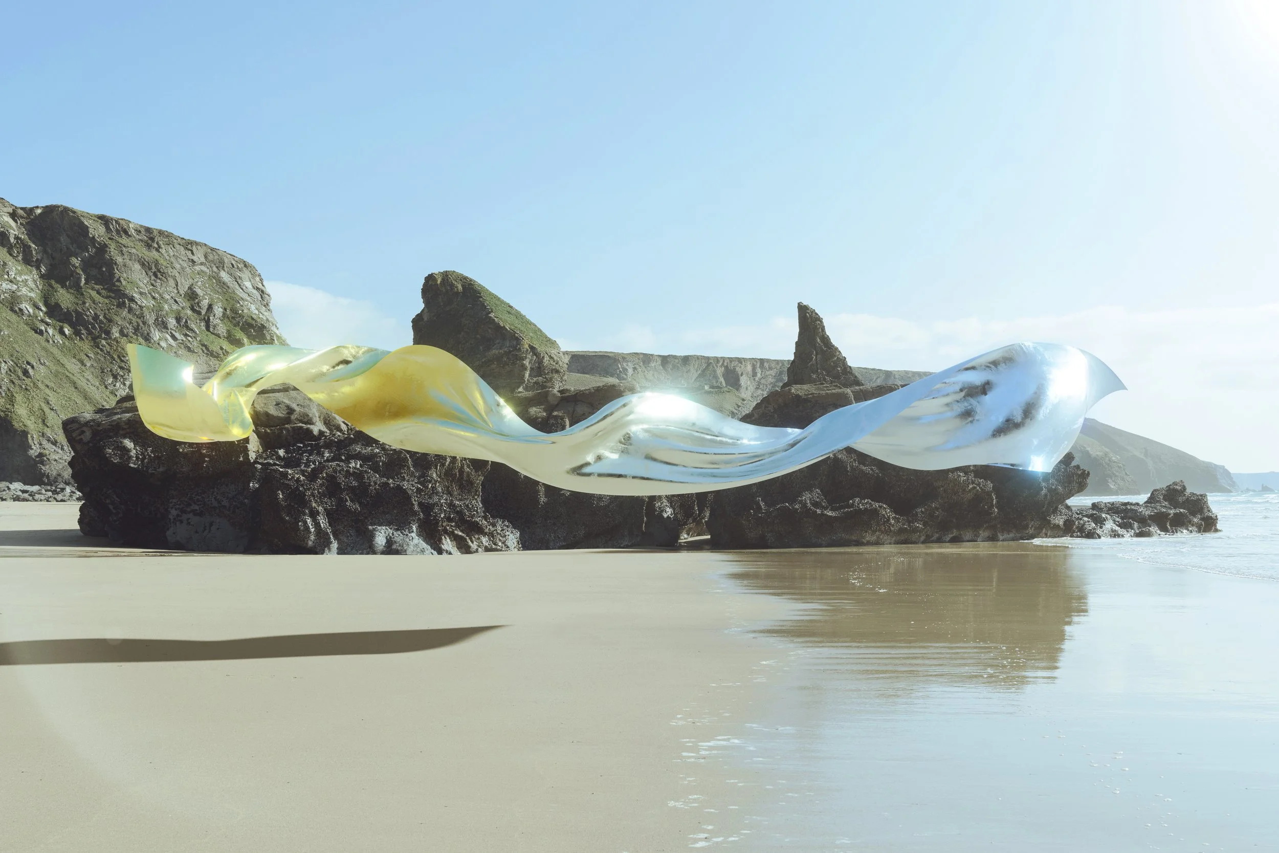 A large, abstract, glass sculpture with yellow and clear sections rests on rocks at a beach, with cliffs and blue sky in the background.