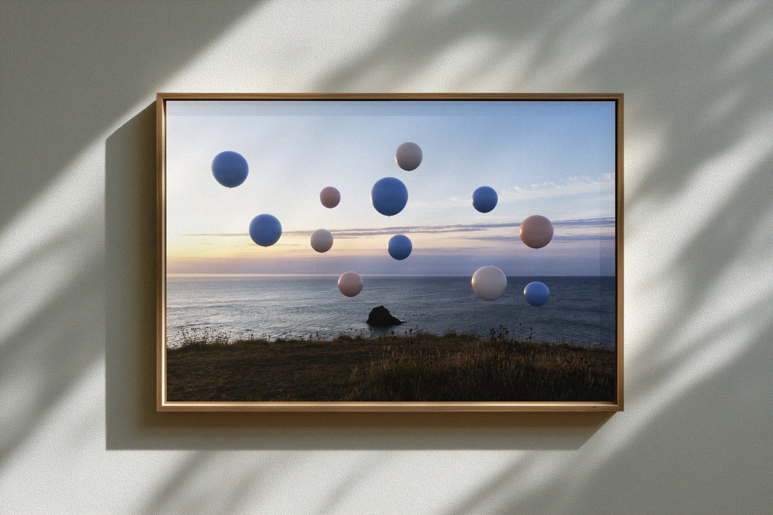 A framed picture of a coastal scene at sunset with multiple floating spheres in blue, white, and pink colors, creating an abstract art composition.