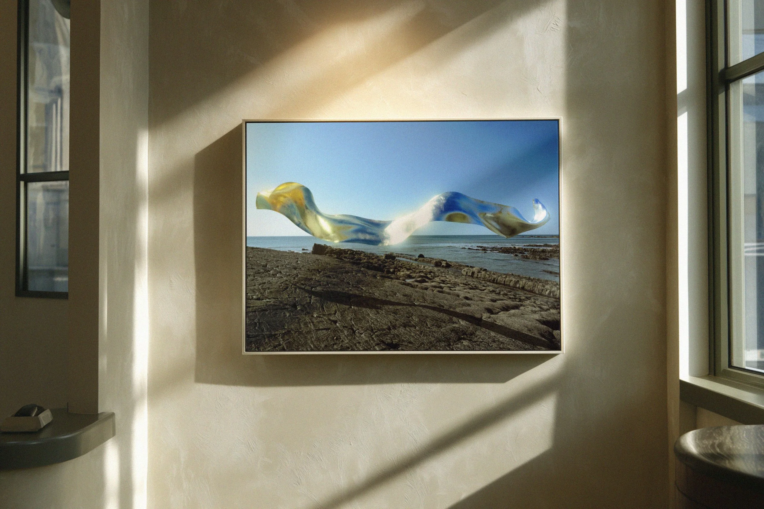 Framed digital artwork of a beach scene with a swirling, abstract, metallic shape floating above the shore, hung on a light-colored wall near windows with sunlight casting shadows.