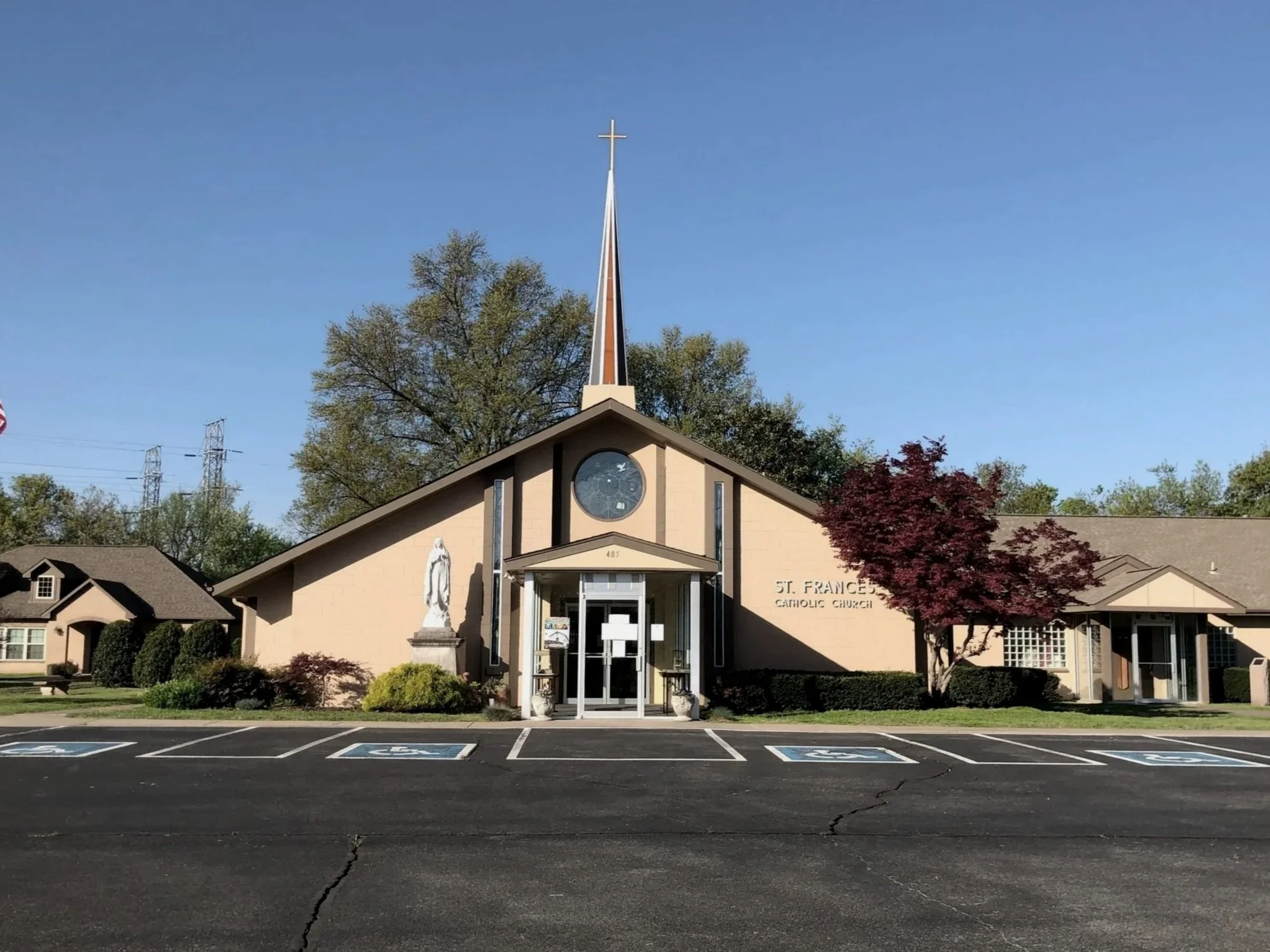 Front view of St. Frances Catholic Church building with parking lot in foreground, featuring four handicapped parking spaces, a statue of a religious figure on the left side, and trees around the building under a clear blue sky.