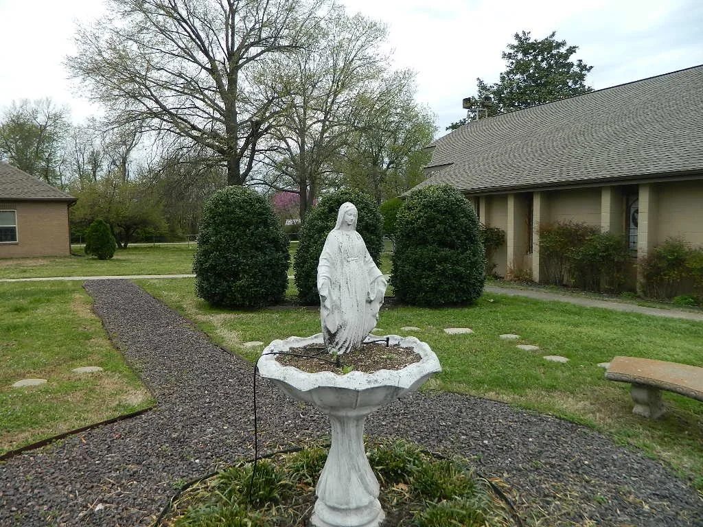 Statue of Jesus Christ standing in a garden fountain, surrounded by trimmed bushes and a gravel pathway.