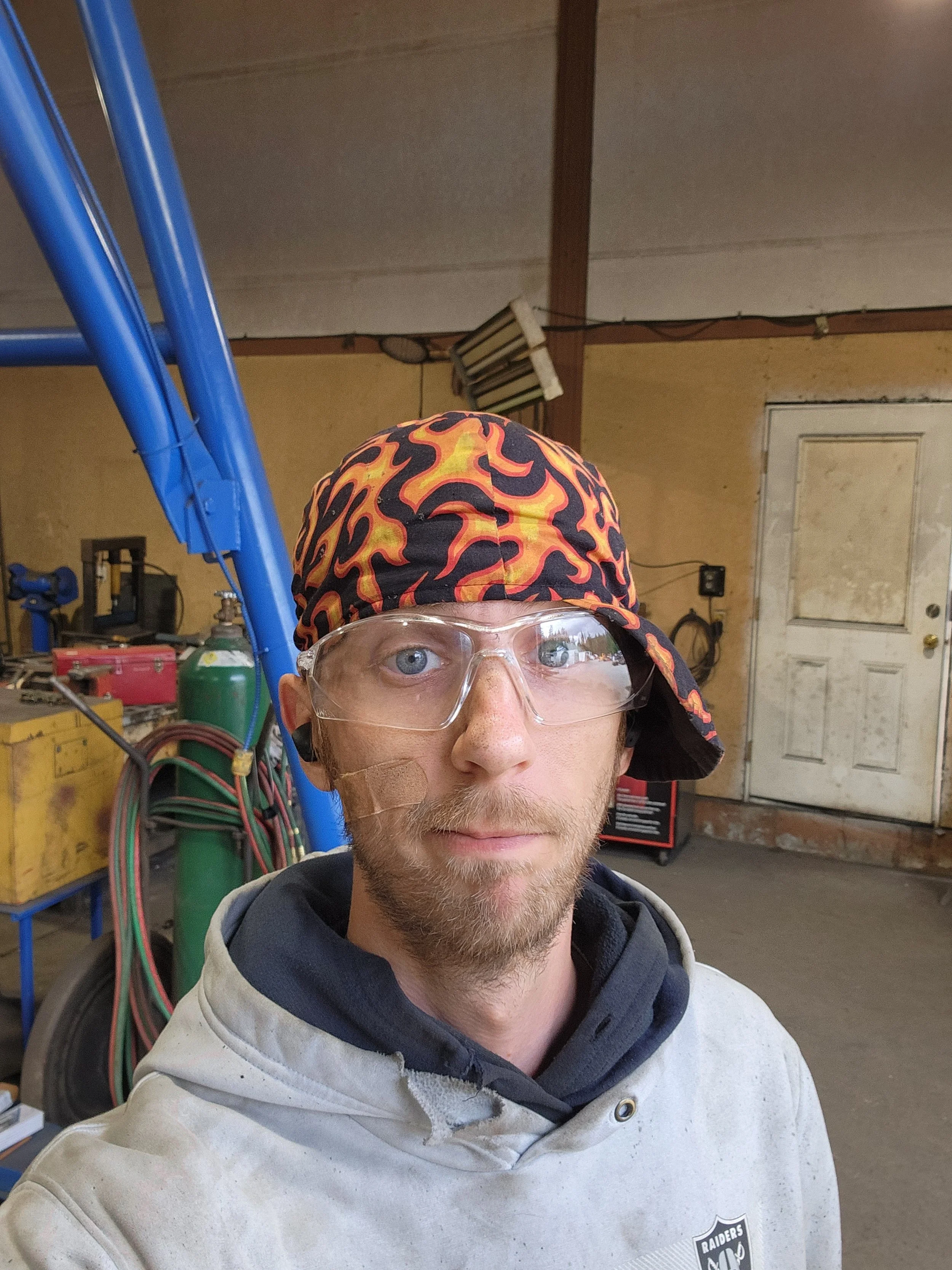 A Welder with safety glasses wearing a cap and hoodie in a workshop, with welding tools and equipment in the background.