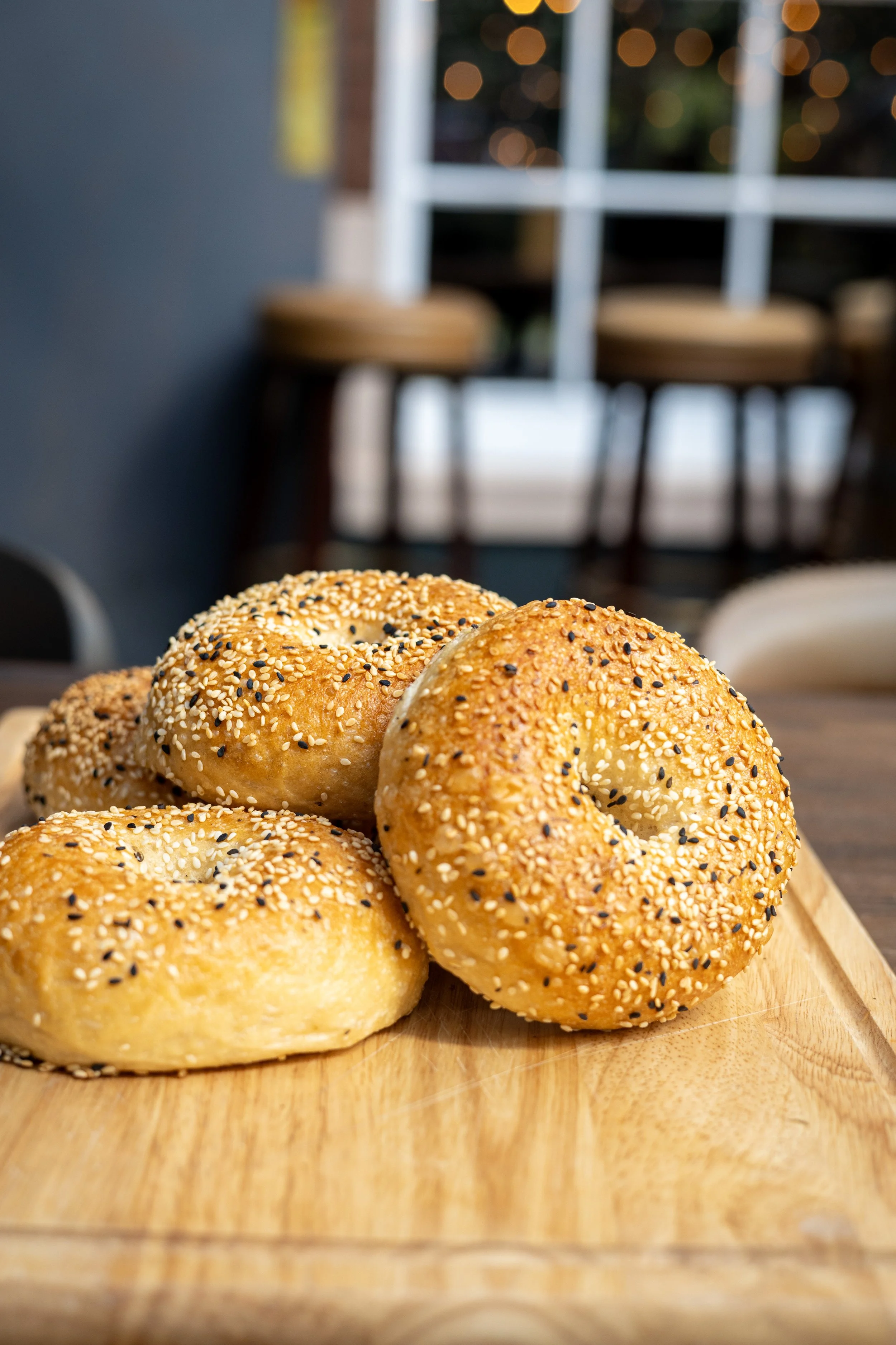 Close-up of sesame and poppy seed bagels on a wooden board with a blurred dining area in the background.