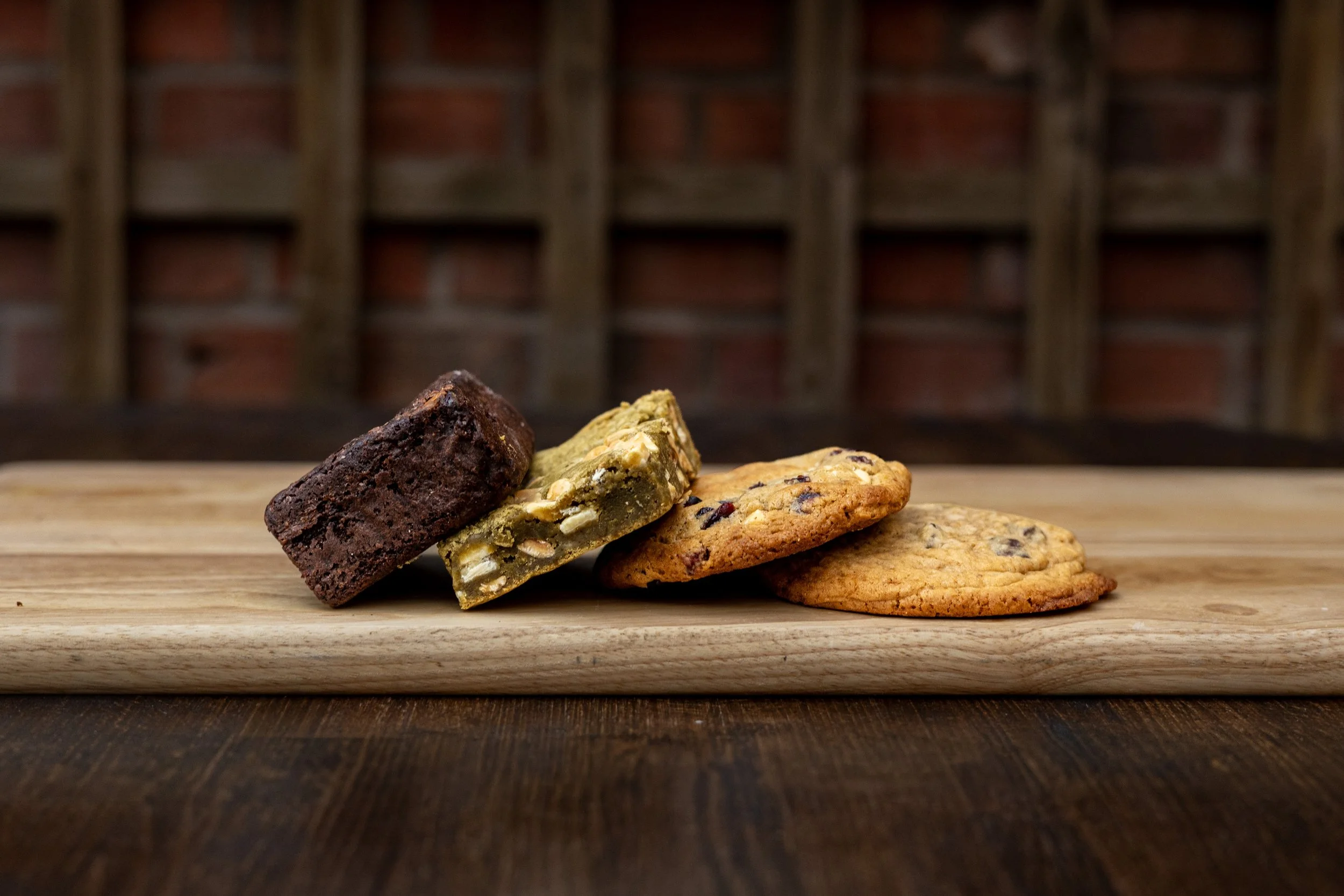 A wooden surface with four cookies and a brownie, with a blurred brick wall in the background.
