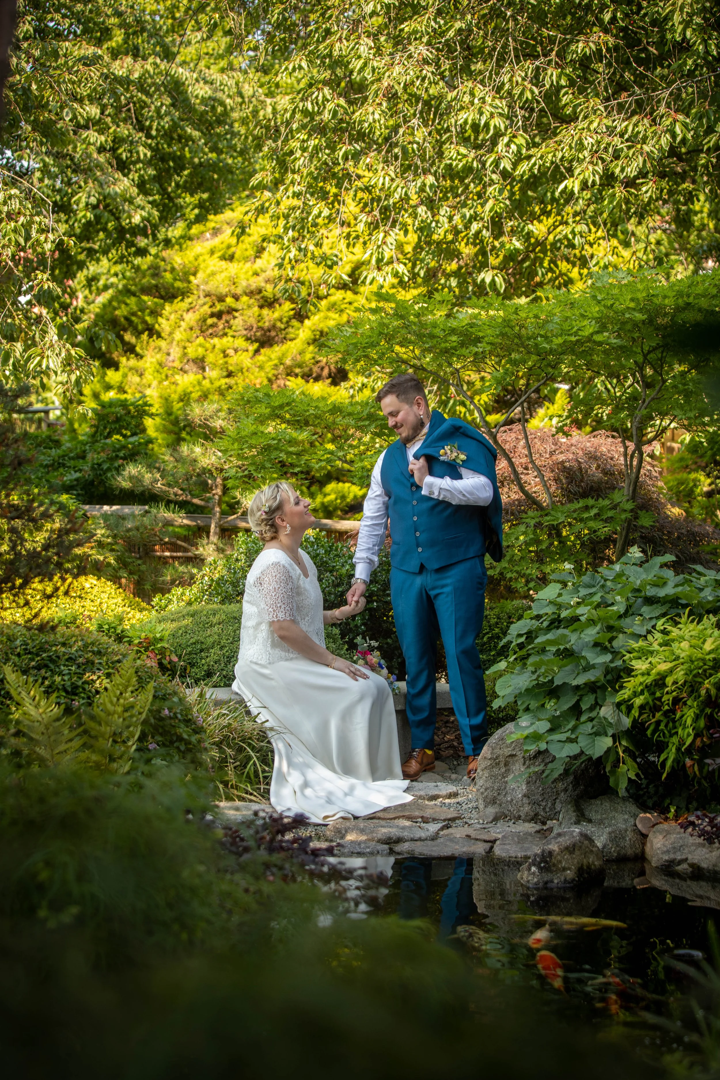 Un couple lors d'un mariage dans un jardin verdoyant, où la femme est assise sur un banc et le mari se tient devant elle, se tenant la main, dans un cadre naturel avec un petit étang et des poissons.