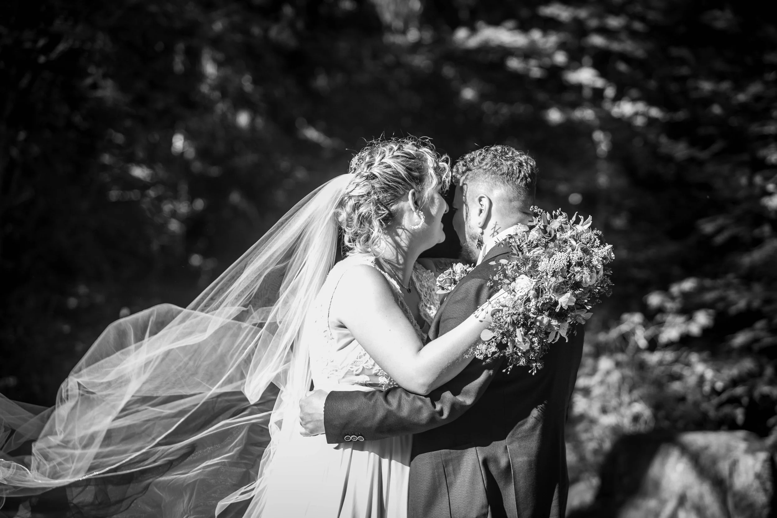Un couple de mariés en plein air, la mariée avec une robe blanche et un voile, et le marié en costume sombre, s'enlacent tendrement, la mariée tenant un bouquet de fleurs. La scène est en noir et blanc, avec des arbres en arrière-plan.