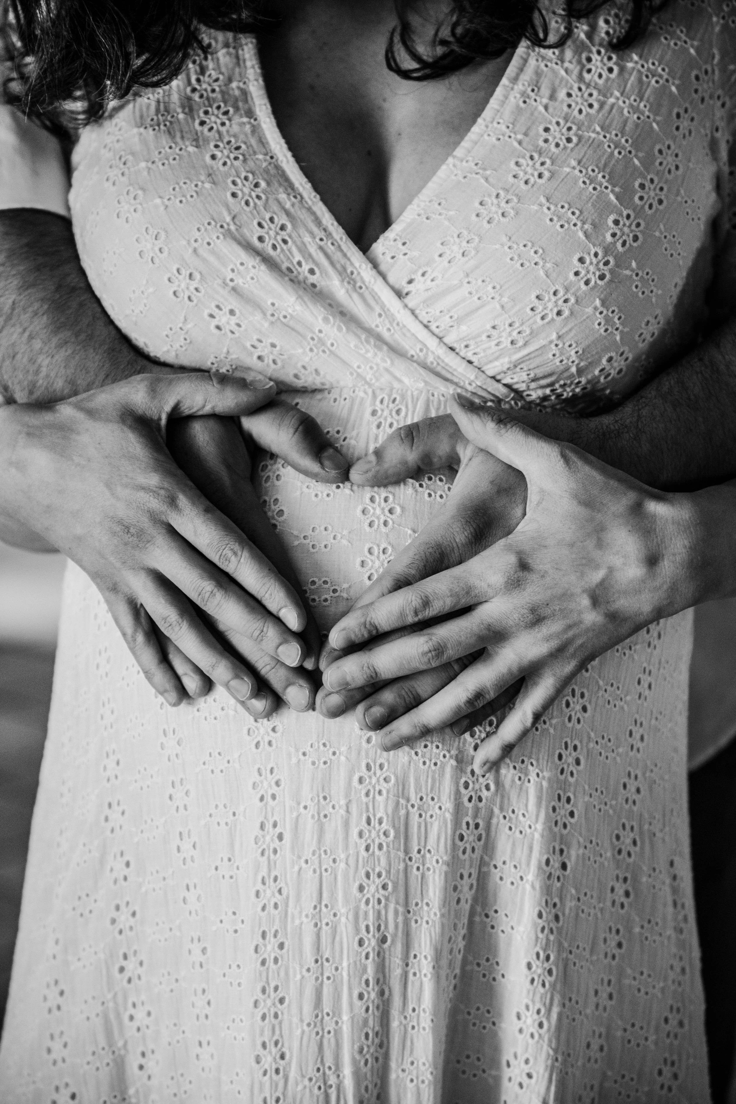 Une femme porte une robe blanche en dentelle avec des motifs de fleurs, et plusieurs mains masculines forment un cœur au niveau du ventre, symbolisant la grossesse et l'amour.
