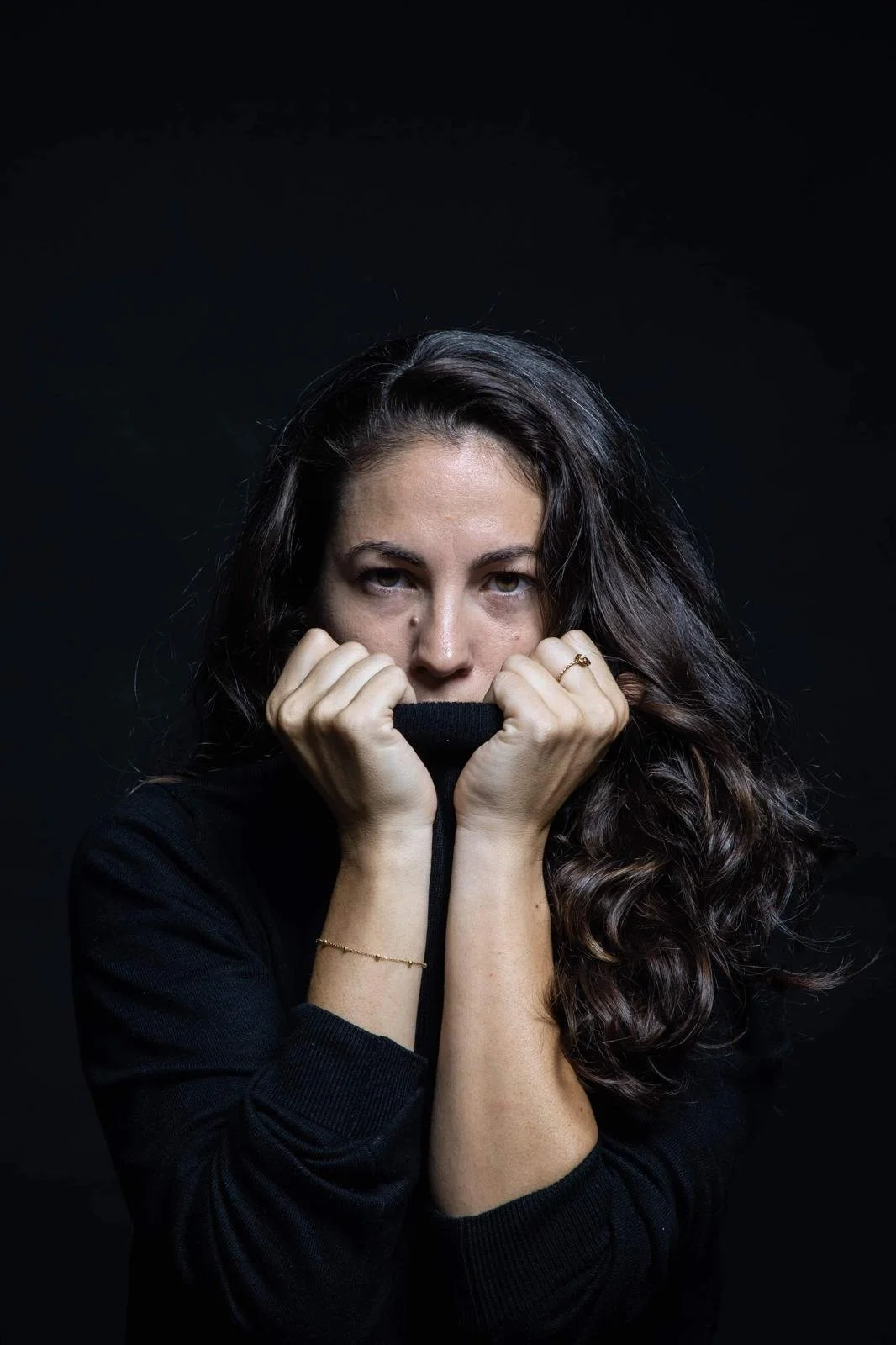 Une femme à cheveux longs et bouclés, portant un sweat noir, se cache partiellement derrière un col de vêtement noire, regardant fixement l'objectif avec une expression sérieuse, contre un fond noir.