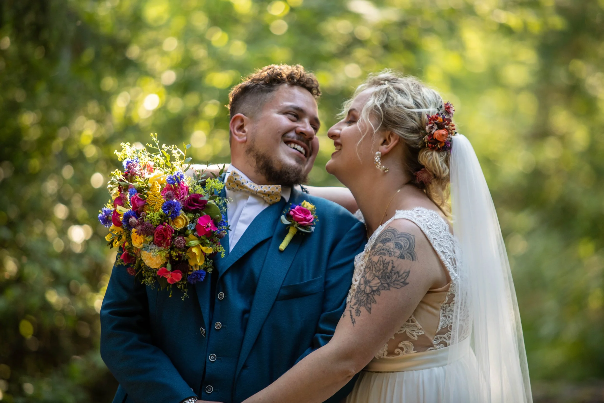 Deux personnes en tenues de mariage, dans un environnement forestier, souriant et se regardant tendrement, avec un bouquet de fleurs colorées.