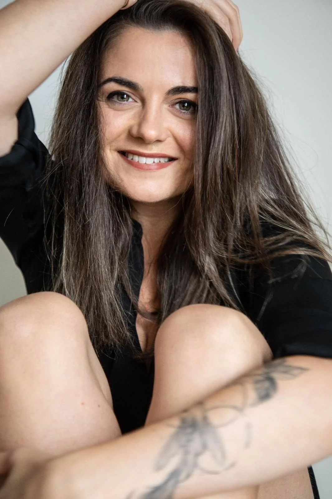 Une femme souriante avec des cheveux bruns, assise avec les genoux pliés, portant un vêtement noir, montrant un tatouage sur le bras, dans un cadre intérieur