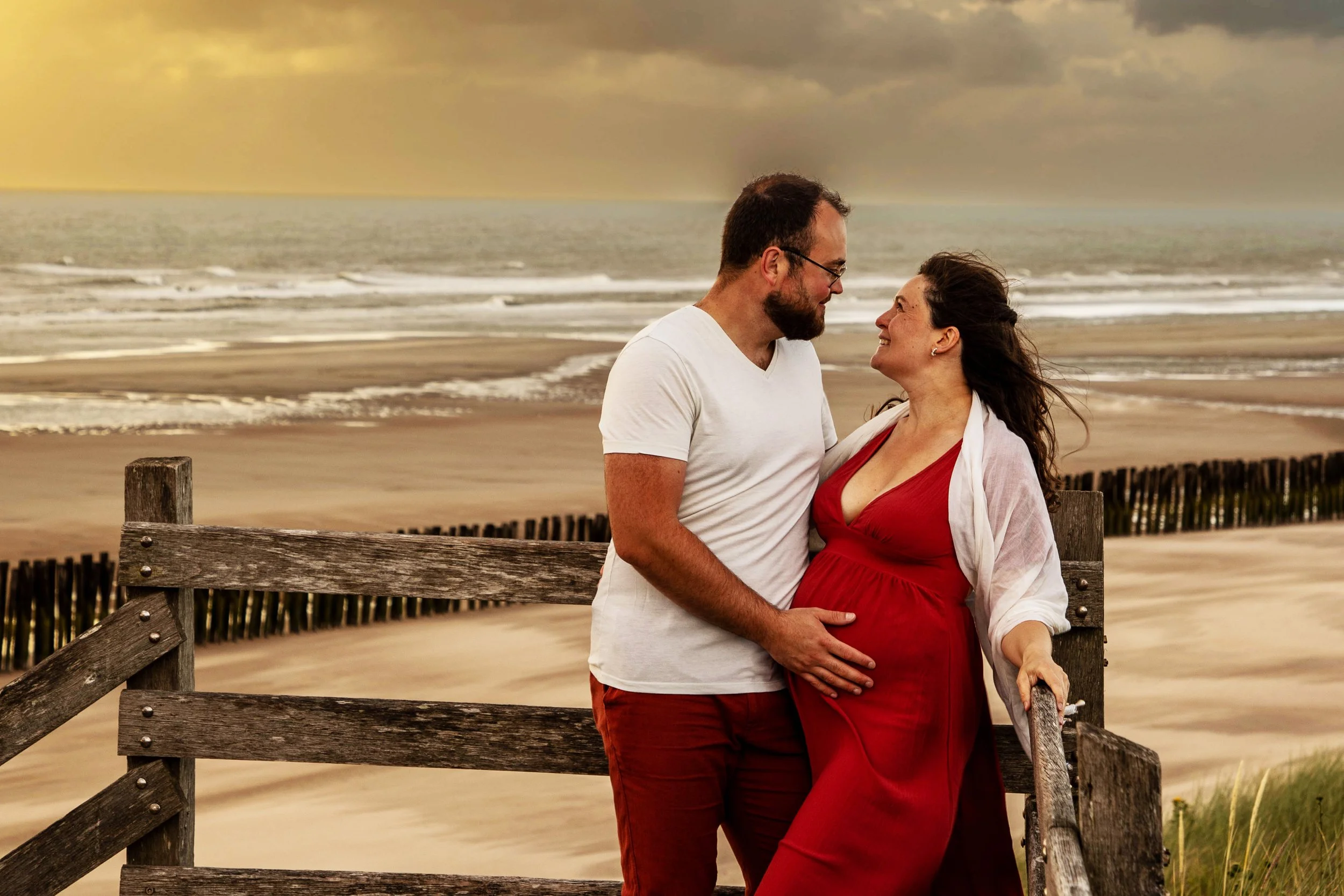 Un couple regarde tendrement en se tenant par la main sur un pont en bois au bord de la plage au coucher du soleil.
