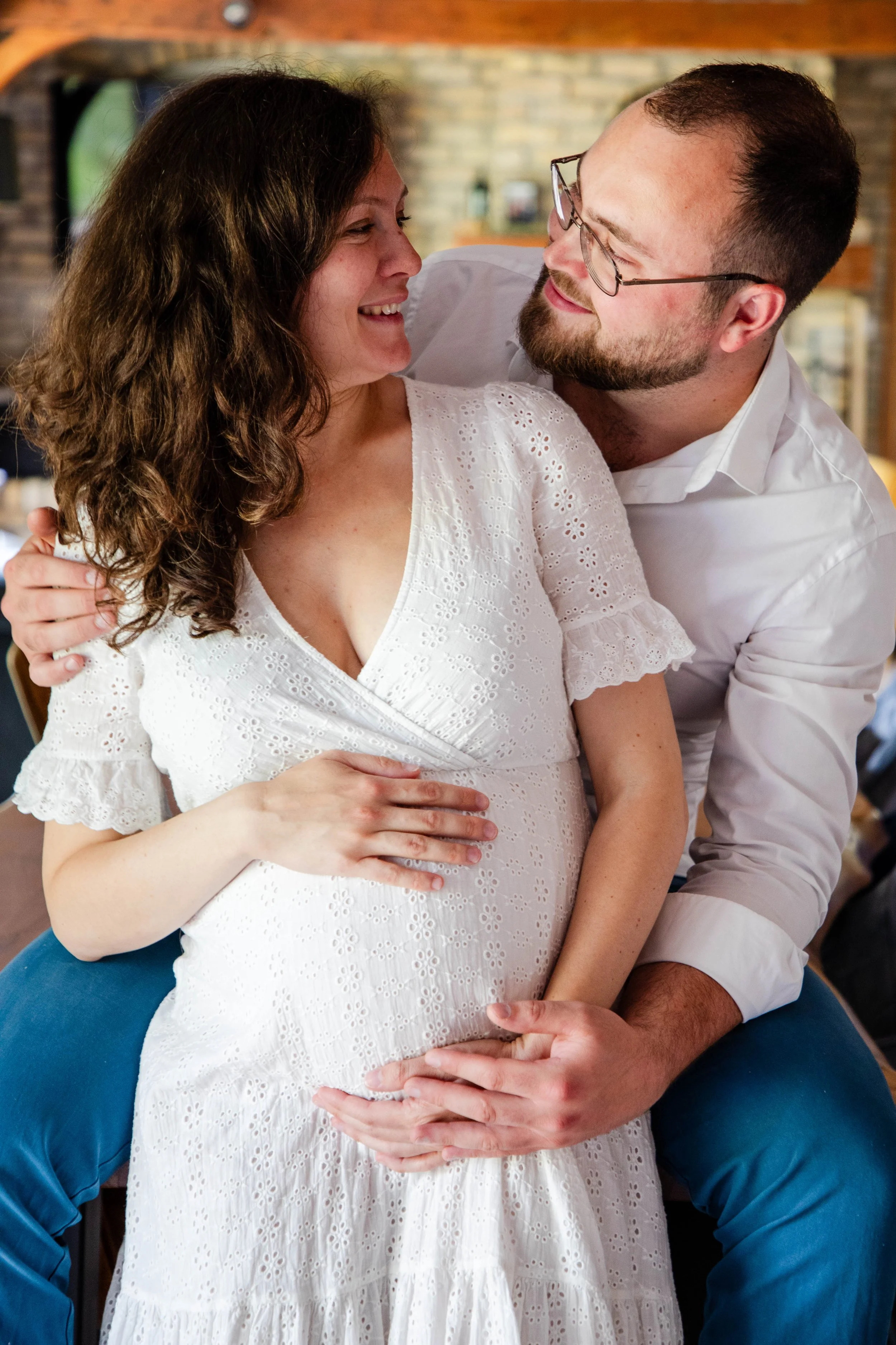 Un couple souriant, le homme touchant le ventre de la femme enceinte, dans un cadre intérieur chaleureux.