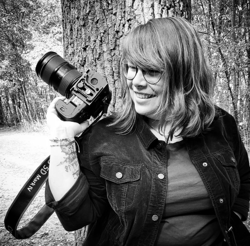 Une femme avec des lunettes tient un appareil photo reflex numérique dans un parc forestier.