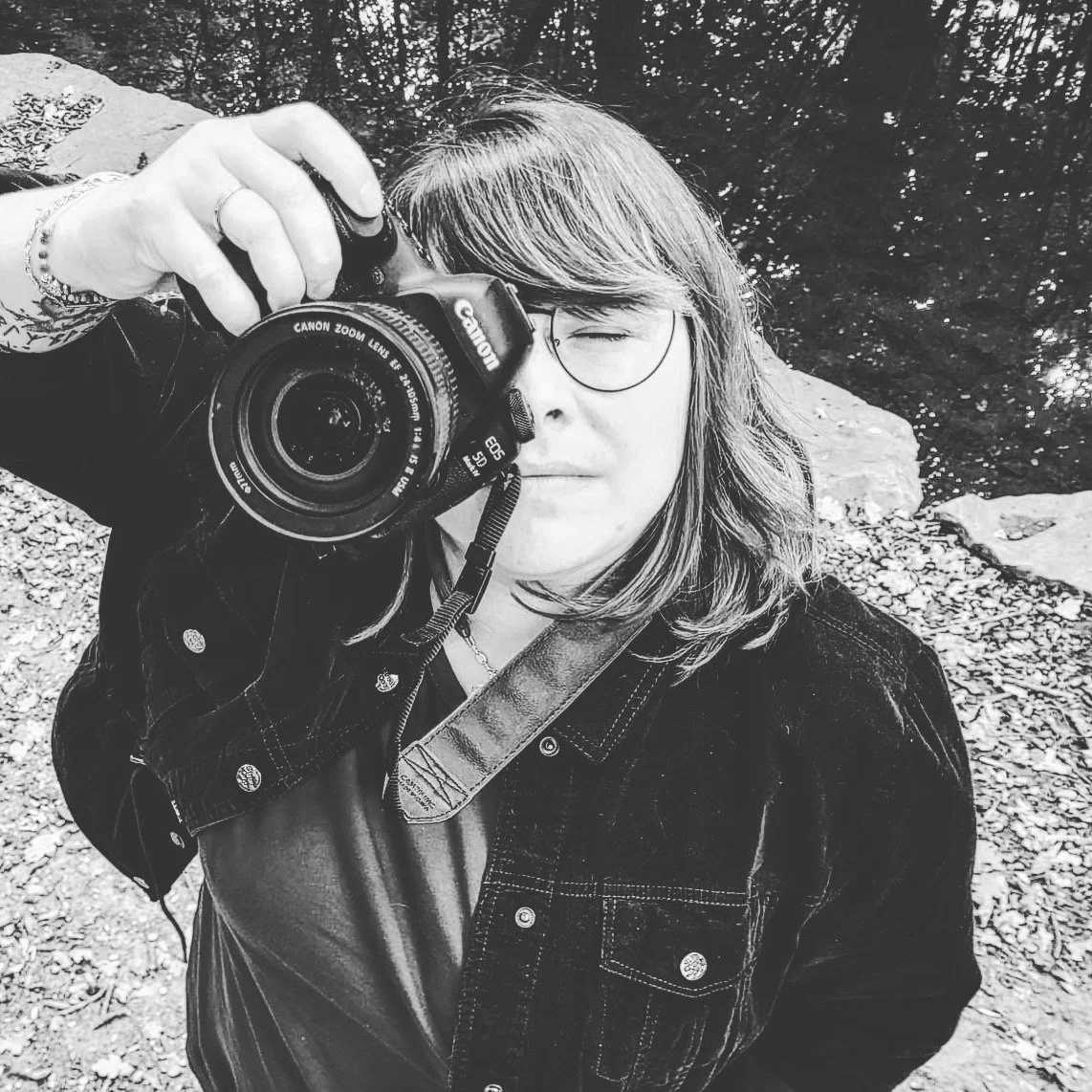 Une personne avec des cheveux courts et une veste en jean, tenant un appareil photo Canon et prenant une photo d'elle-même dans un environnement naturel avec des rochers et des arbres.
