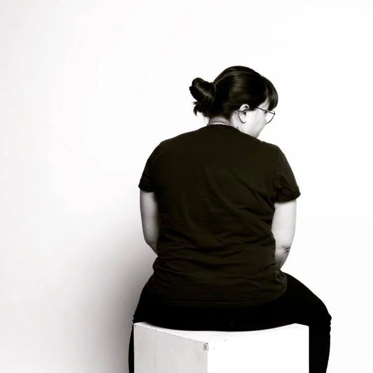 Une femme avec des cheveux foncés noués en chignon, portant des lunettes et un t-shirt noir, vue de dos, assise sur un cube blanc devant un mur blanc.