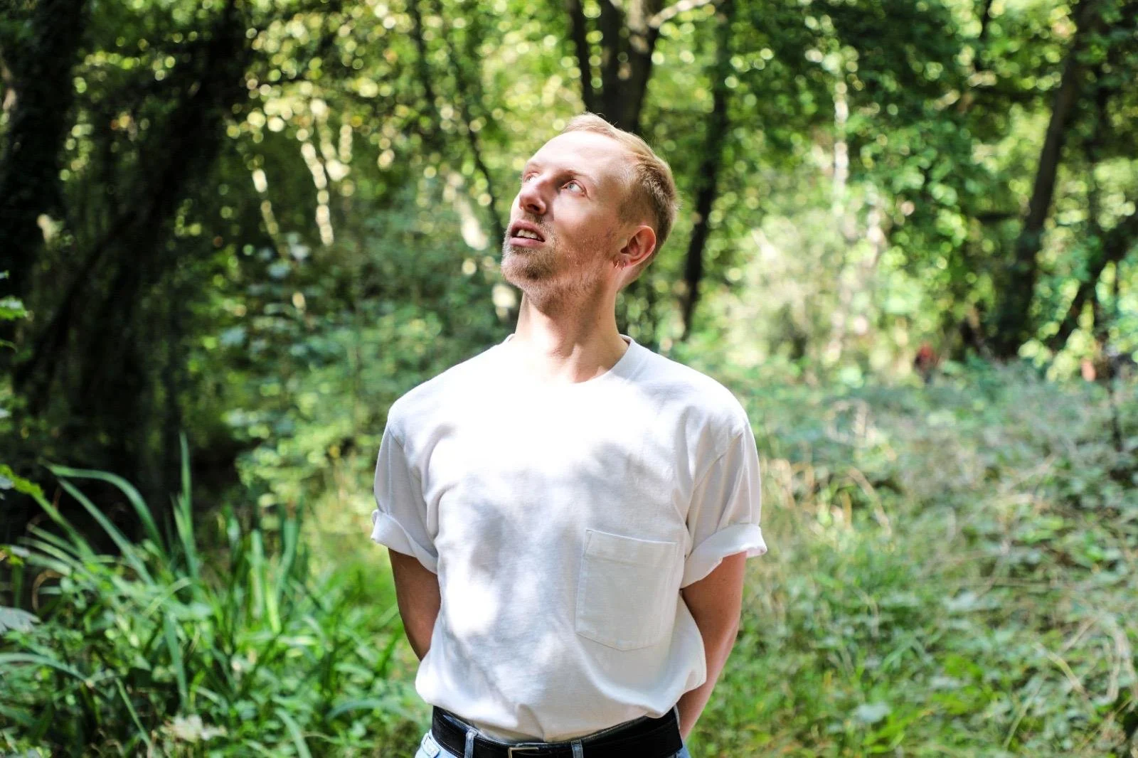 Jeune homme debout dans une forêt verdoyante, portant un t-shirt blanc avec les manches roulées, regardant vers le haut.