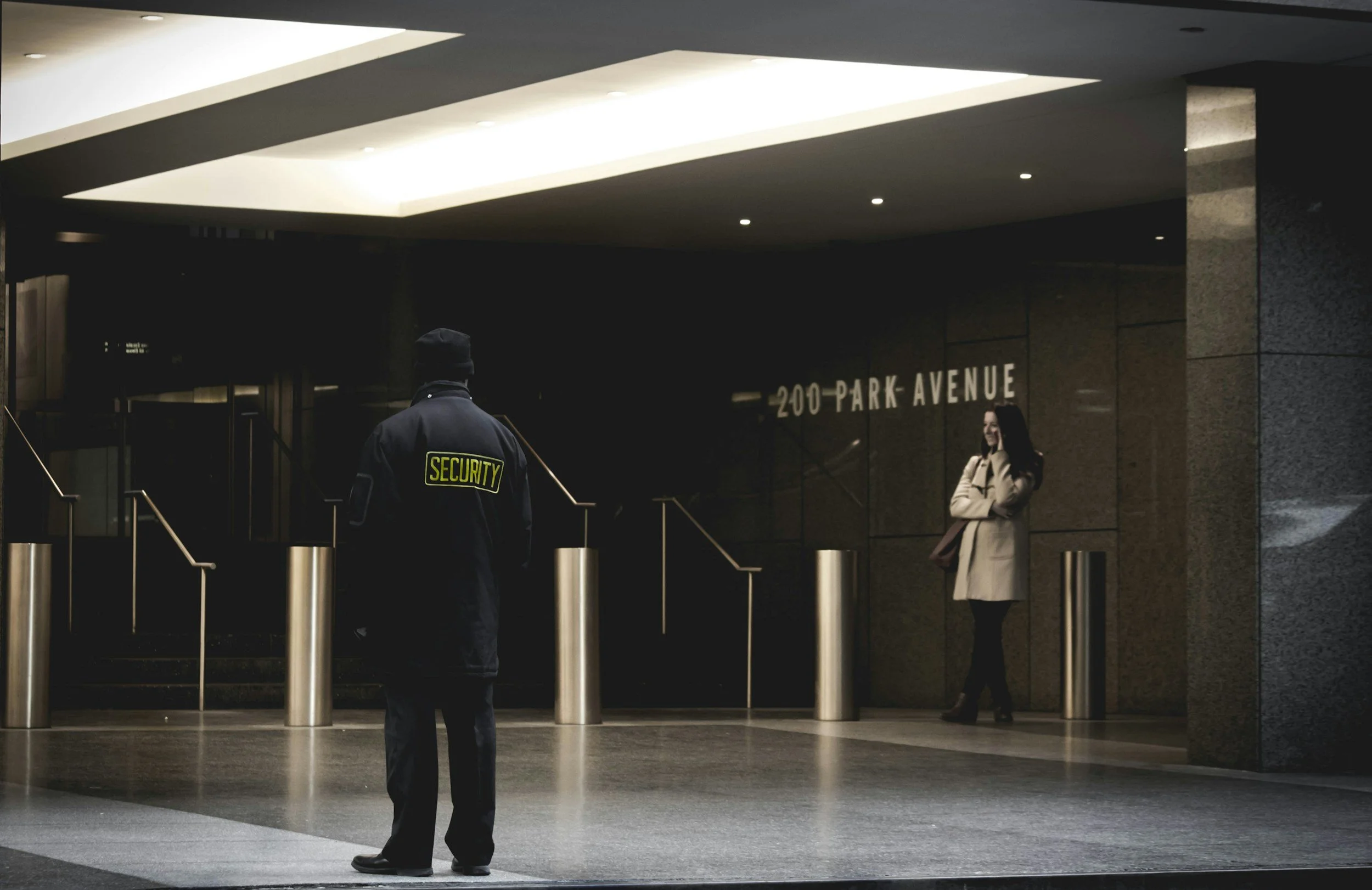 Security guard standing outside a building at 200 Park Avenue, with a woman in a beige coat talking on her phone in the background.