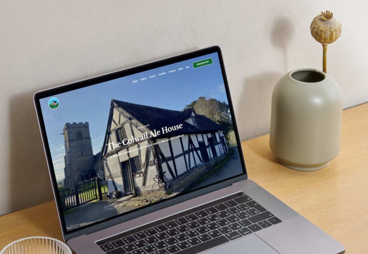 A laptop on a wooden desk displaying a website about The Colwall Ale House, with an old historic building and church tower in the background, next to a beige vase with dried poppy seed head, and a small container on the desk.