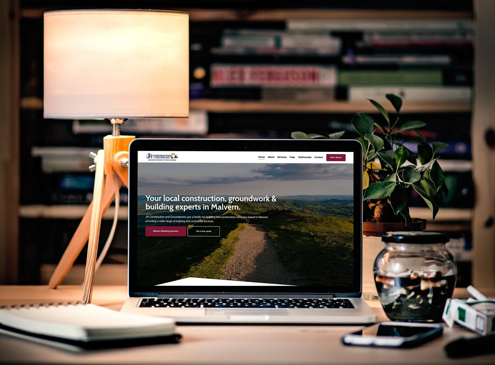 Laptop on a wooden desk displaying a construction company's website with a scenic landscape, a potted plant, a lamp, and various office supplies around it.