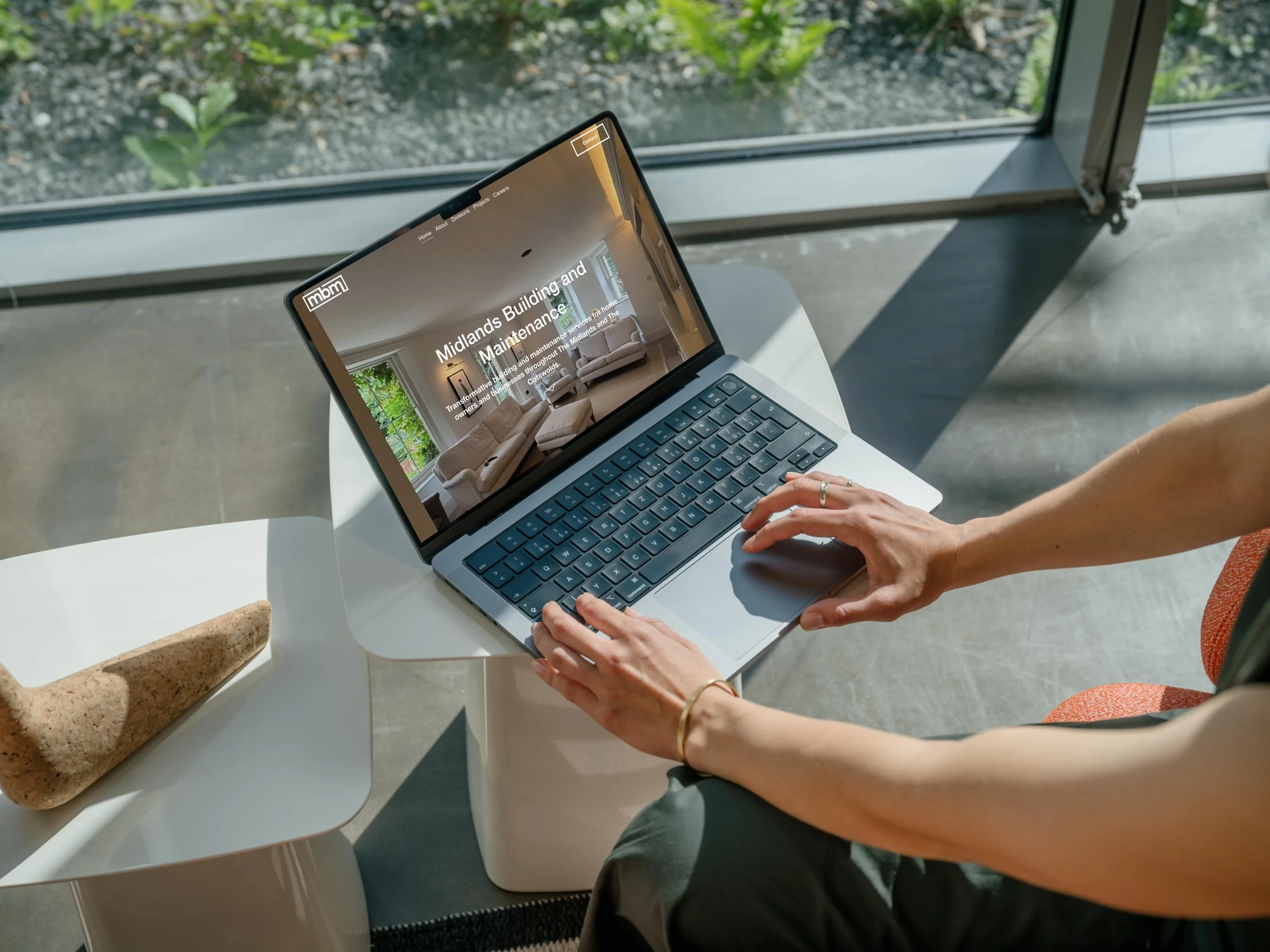 A person using a laptop on a white table near a large window, with a view of greenery outside. The laptop screen displays a website about Midlands Building and Maintenance.