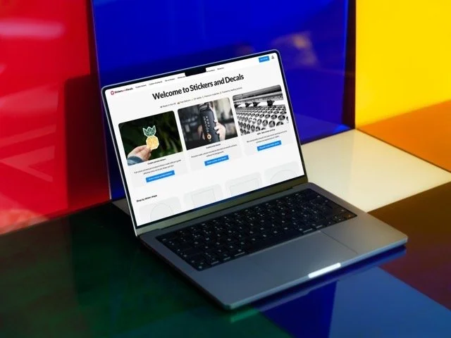 Open laptop on a colorful surface displaying a website titled 'Welcome to Stickers and Decals' with images of a leaf sticker, a marker, and typewriter keys.