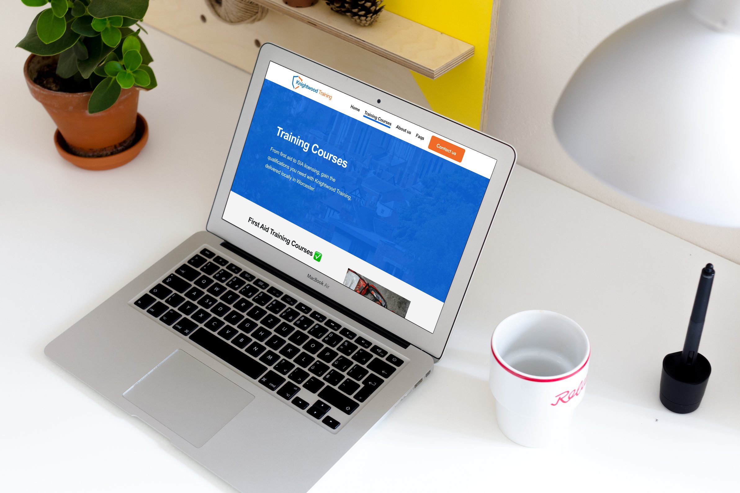 A silver MacBook Air laptop on a white desk displaying a website for Knigthwood Training about training courses. Next to the laptop is a white cup with red stripes and a black straw, a potted plant with green leaves, a pen, and a cup with the Red Lobster logo. A yellow and wood-colored shelf is in the background.