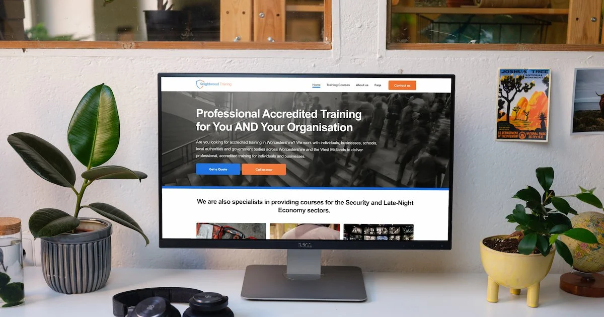 Computer monitor displaying a website for professional accredited training, on a white desk with potted plants, headphones, and framed artwork on the wall.