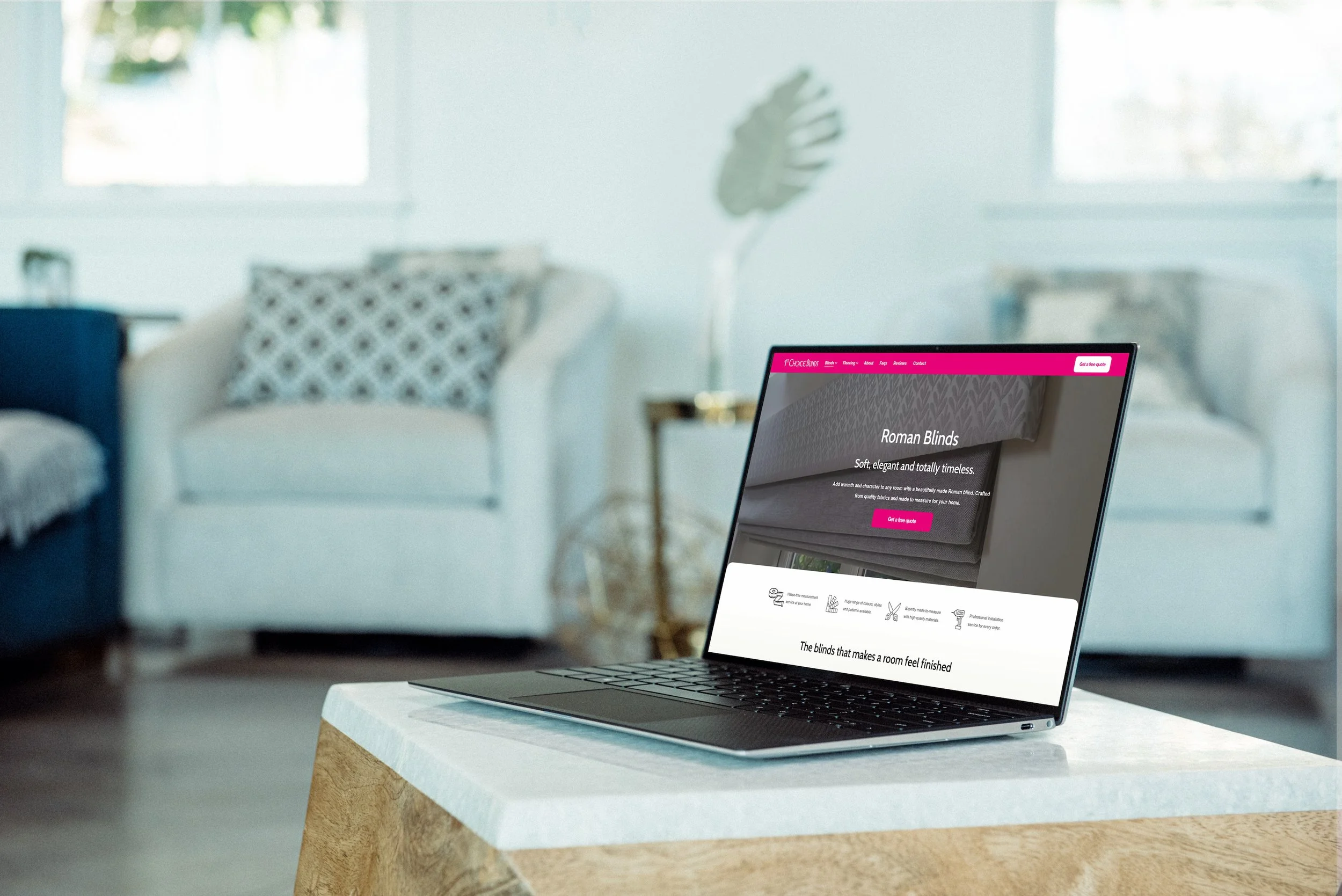 Laptop on a white and wooden table displaying a website about Roman blinds in a bright room with large windows and blurred interior decor objects.