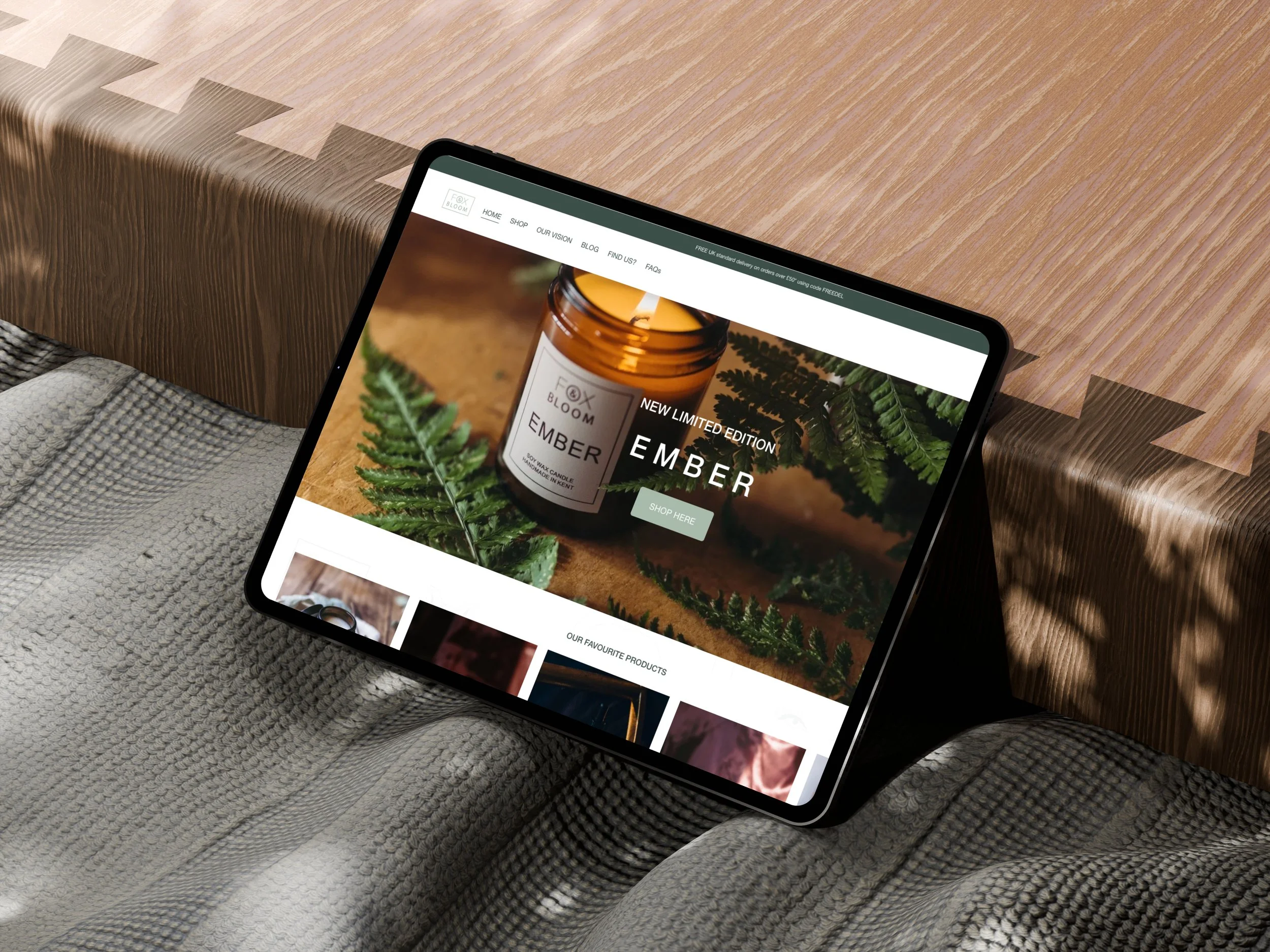 A tablet displaying a website with a product image of a candle labeled 'Ember' and the text 'New Limited Edition Ember.' The tablet is resting on a chair with a textured gray blanket, and there is a wooden surface nearby.